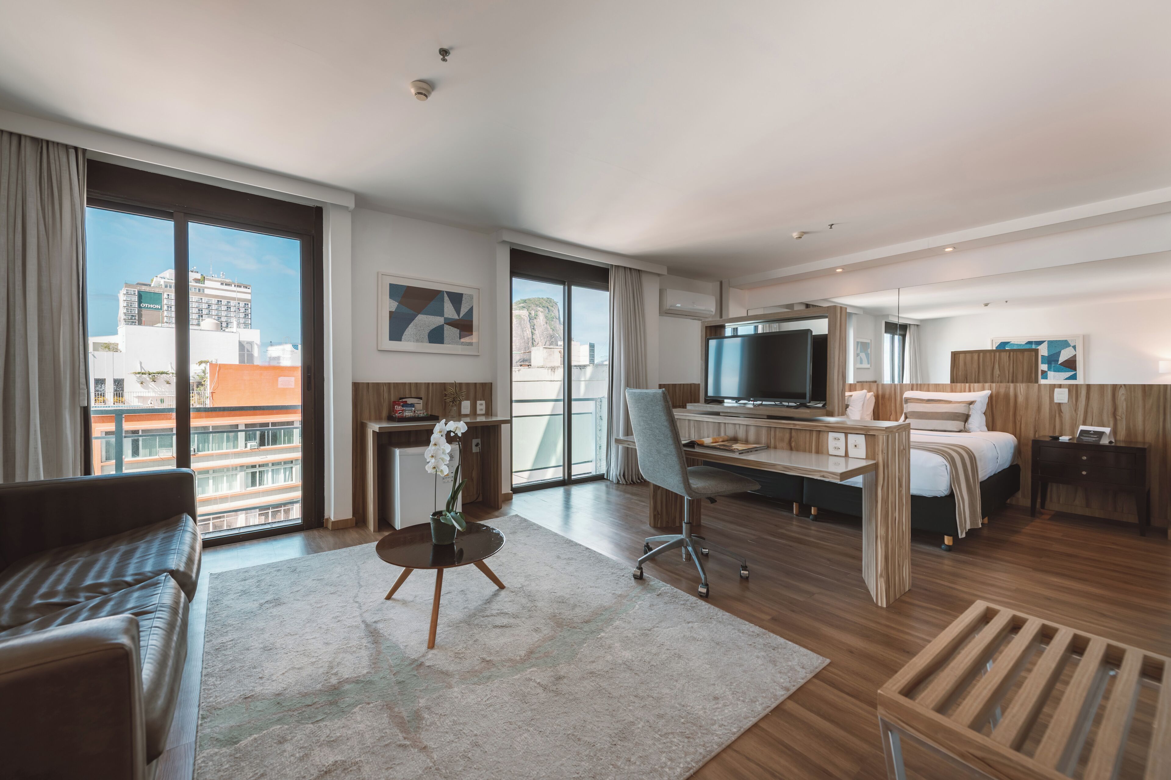 A modern hotel room with a large window, a desk, a bed, a TV, and a seating area.