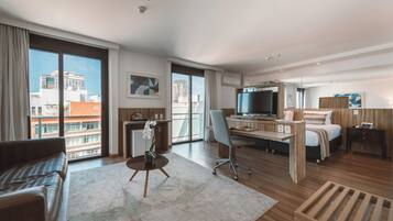 A modern hotel room with a large window, a desk, a bed, a TV, and a seating area.