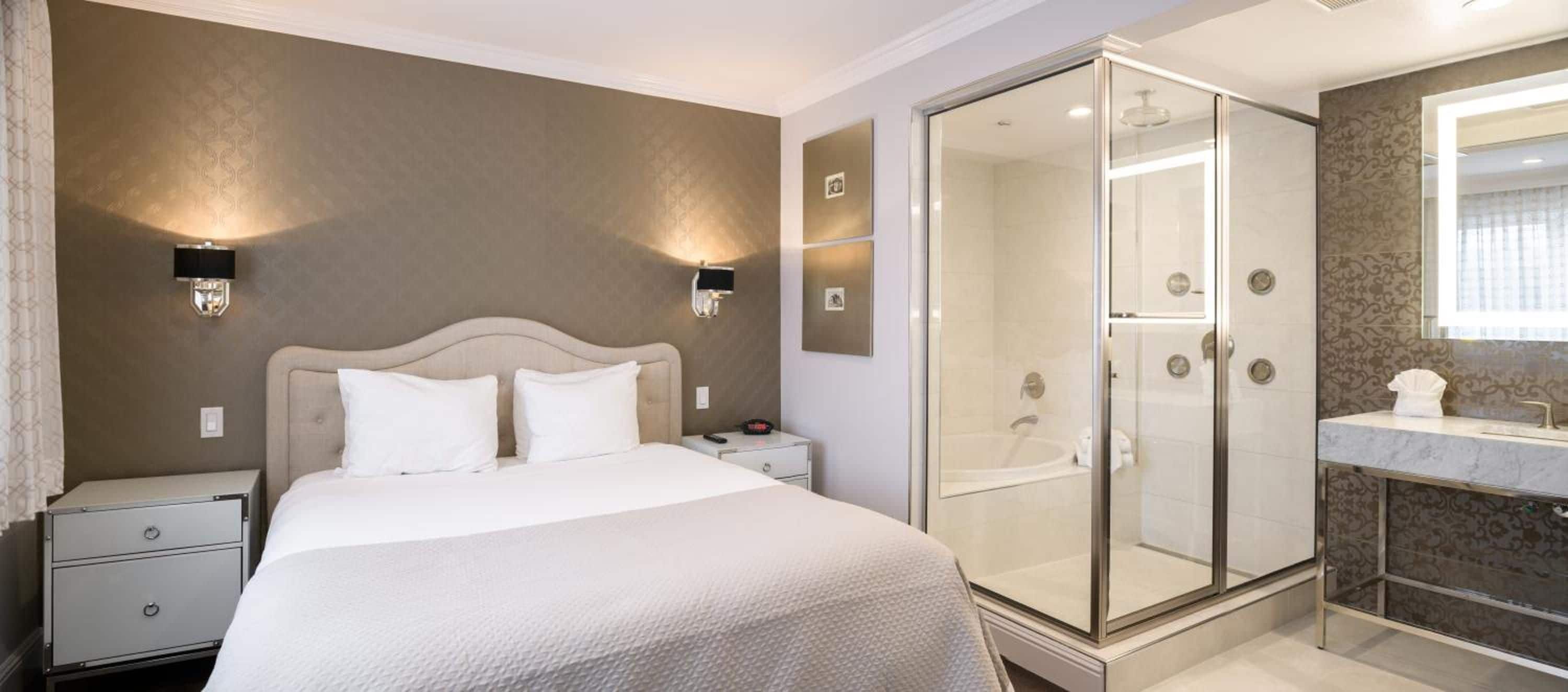 Habitación de hotel con cama, mesitas de noche, ducha a pie y baño con lavamanos y espejo.