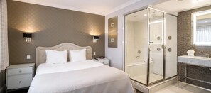 Habitación de hotel con cama, mesitas de noche, ducha a pie y baño con lavamanos y espejo.