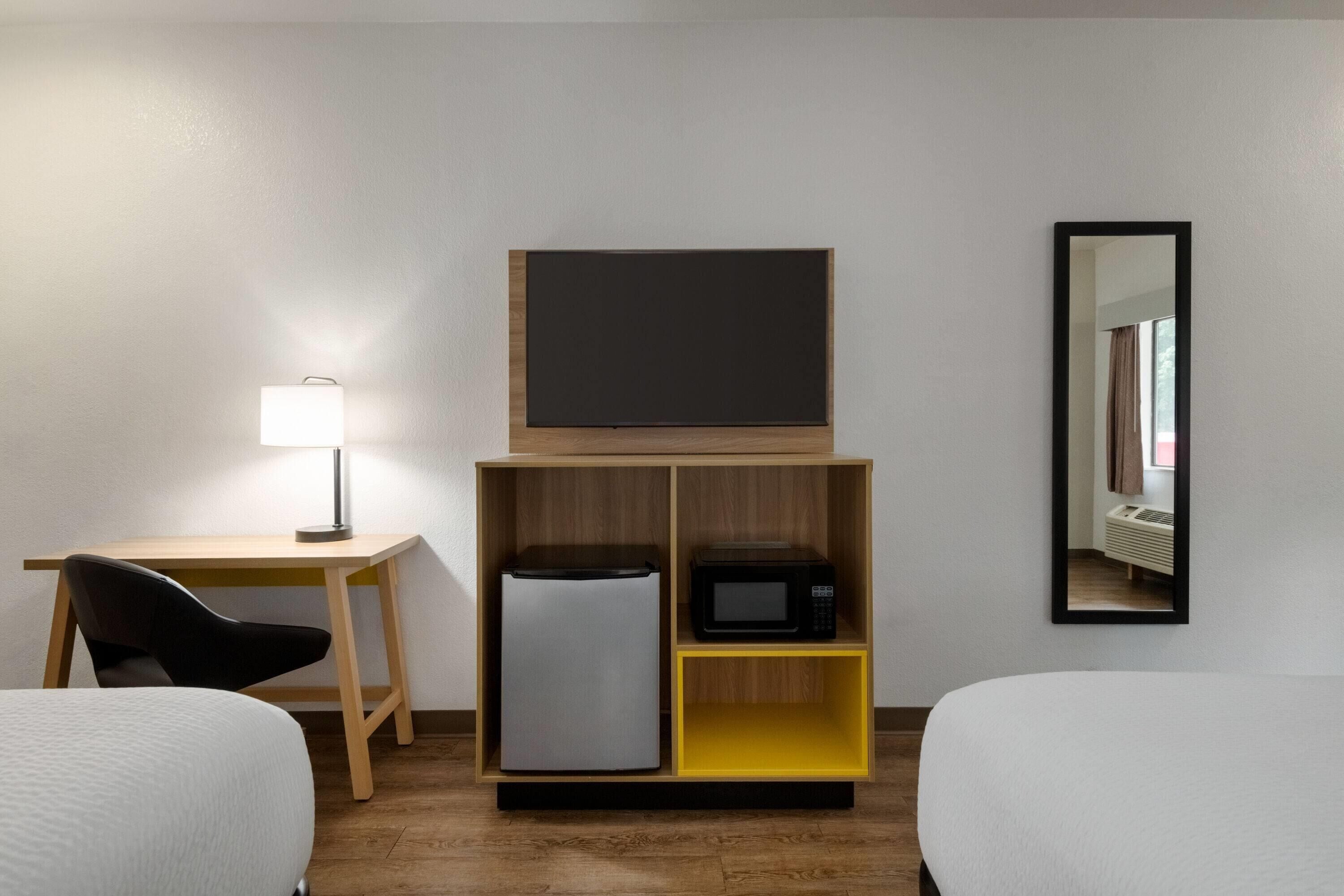 A hotel room with a bed, a desk with a lamp, a refrigerator, a microwave, and a mirror on the wall.