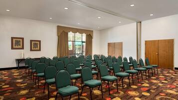 A conference room with rows of green chairs, a patterned carpet, a large window with curtains, and a wooden door.