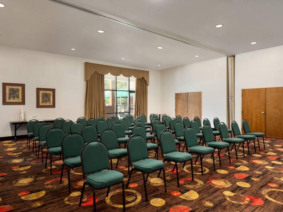A conference room with rows of green chairs, a patterned carpet, a large window with curtains, and a wooden door.