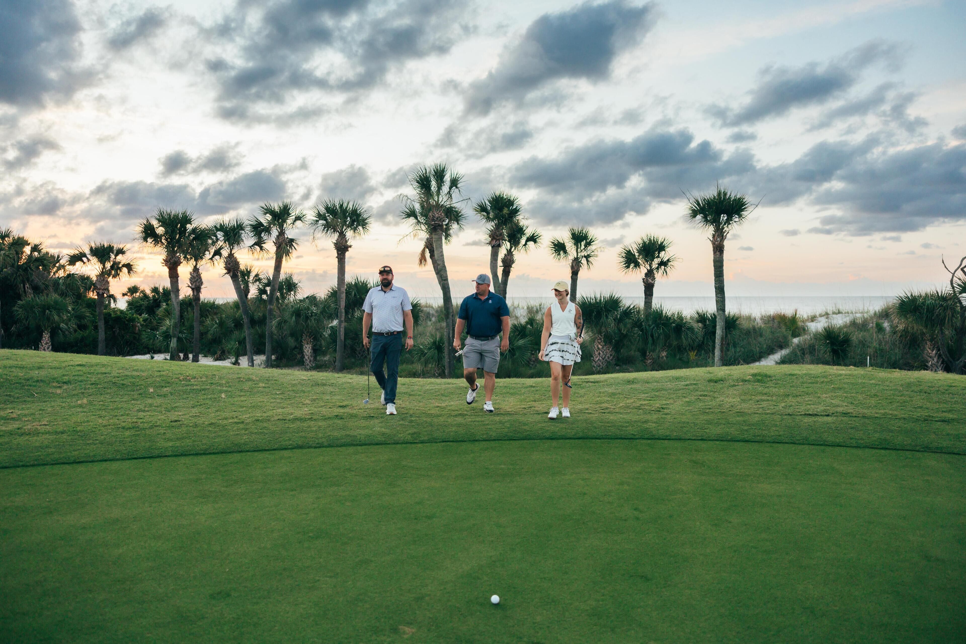 A golf course with three people standing on the green, palm trees in the background, and a cloudy sky.
