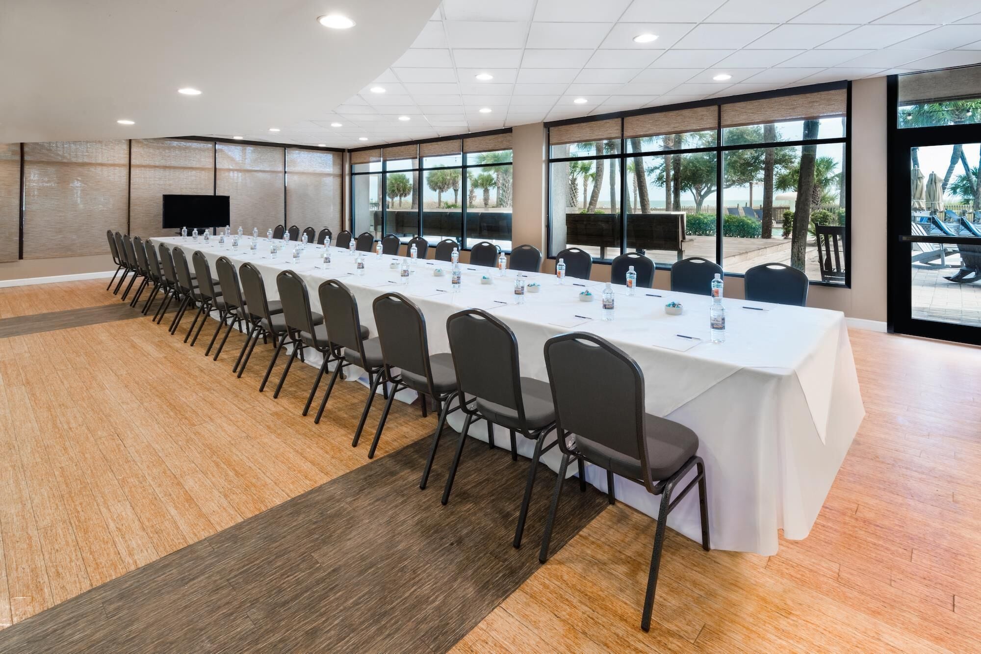 A conference room with a long table, chairs, and large windows offering a view of palm trees and a beach.