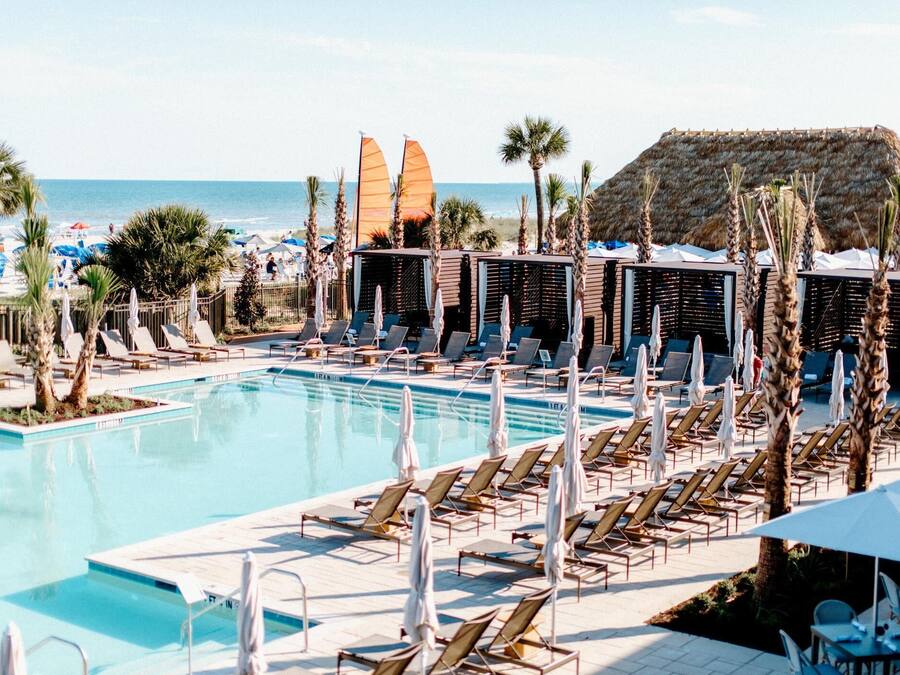 A poolside area with lounge chairs and umbrellas, a clear view of the ocean, and a thatched-roof structure in the background.
