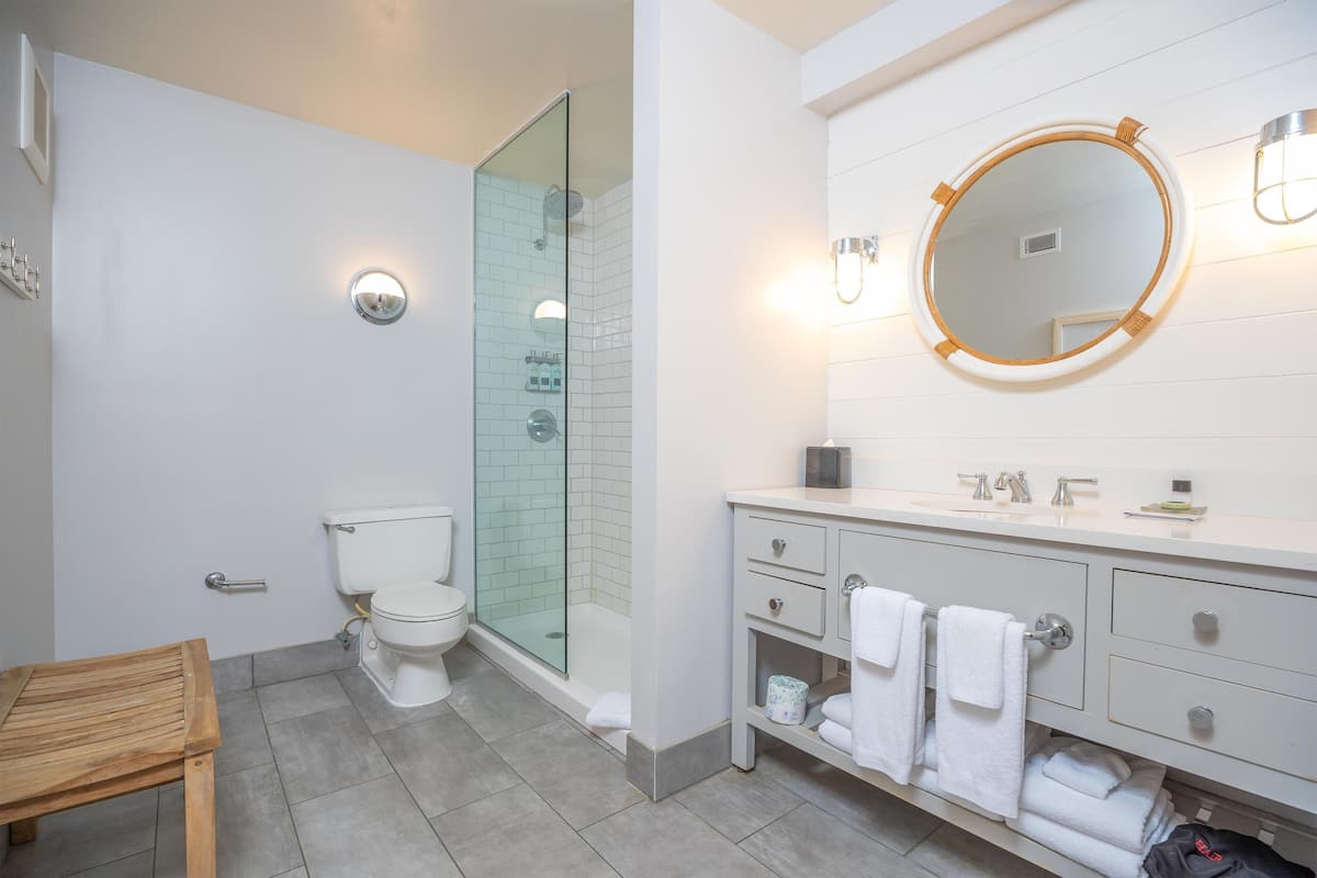 A modern bathroom with a glass-enclosed shower, a white sink with a round mirror, and a wooden bench.