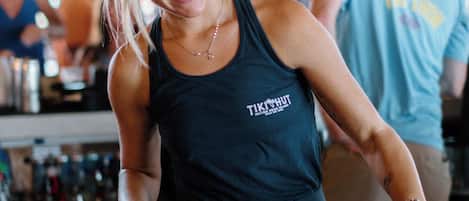 Una persona con una camiseta oscura que muestra un logotipo, de pie en una zona de bar con estantes llenos de botellas y vasos.