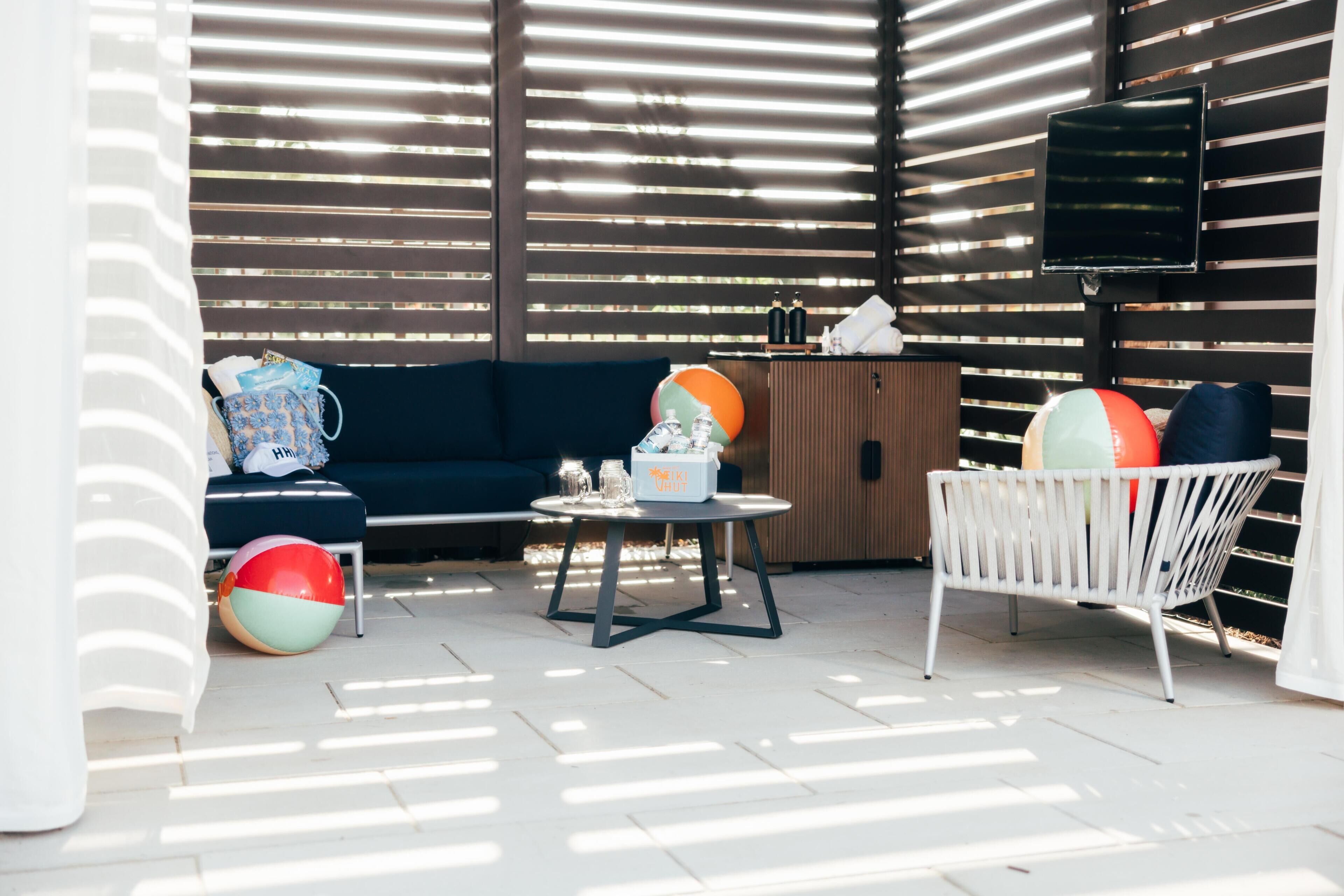 A patio area with a sofa, a round table, and a white chair. There are two beach balls and a small table with bottles and a container.