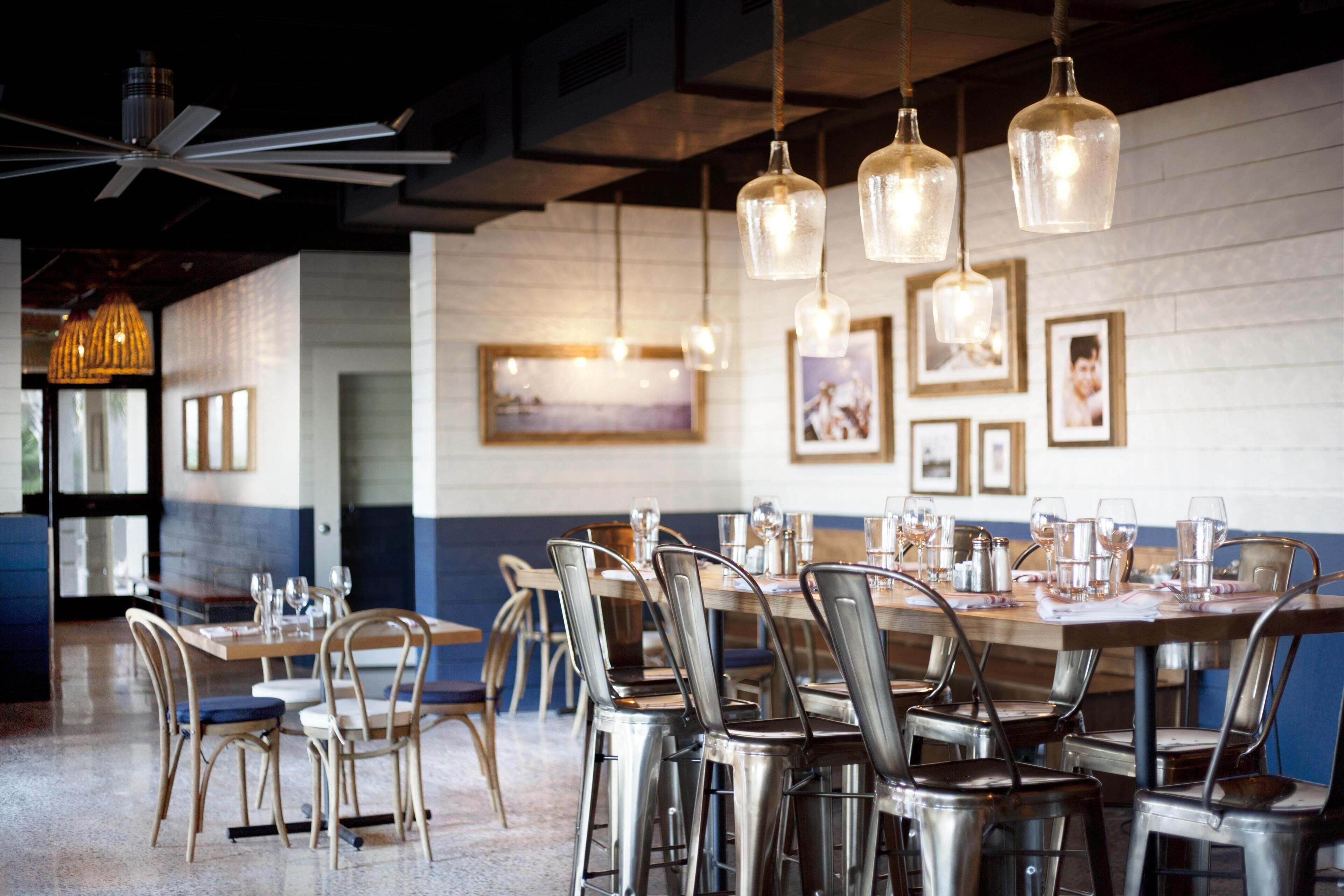 A restaurant with a mix of metal and wooden chairs, a long table set for dining, and hanging pendant lights.