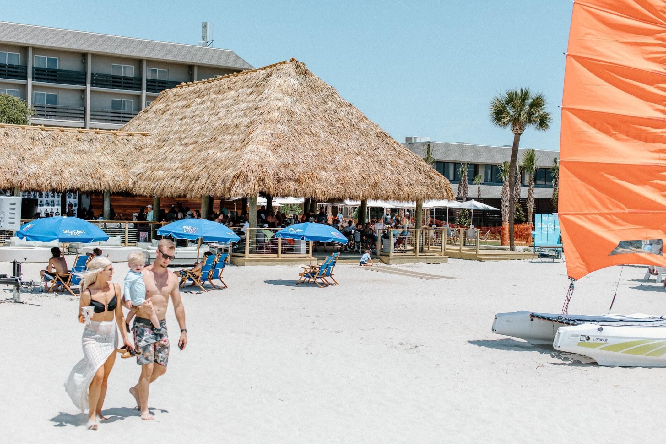A beachside resort with thatched-roof structures, lounge chairs, and a sailboat.