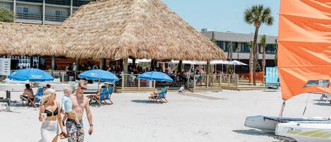 A beachside resort with thatched-roof structures, lounge chairs, and a sailboat.