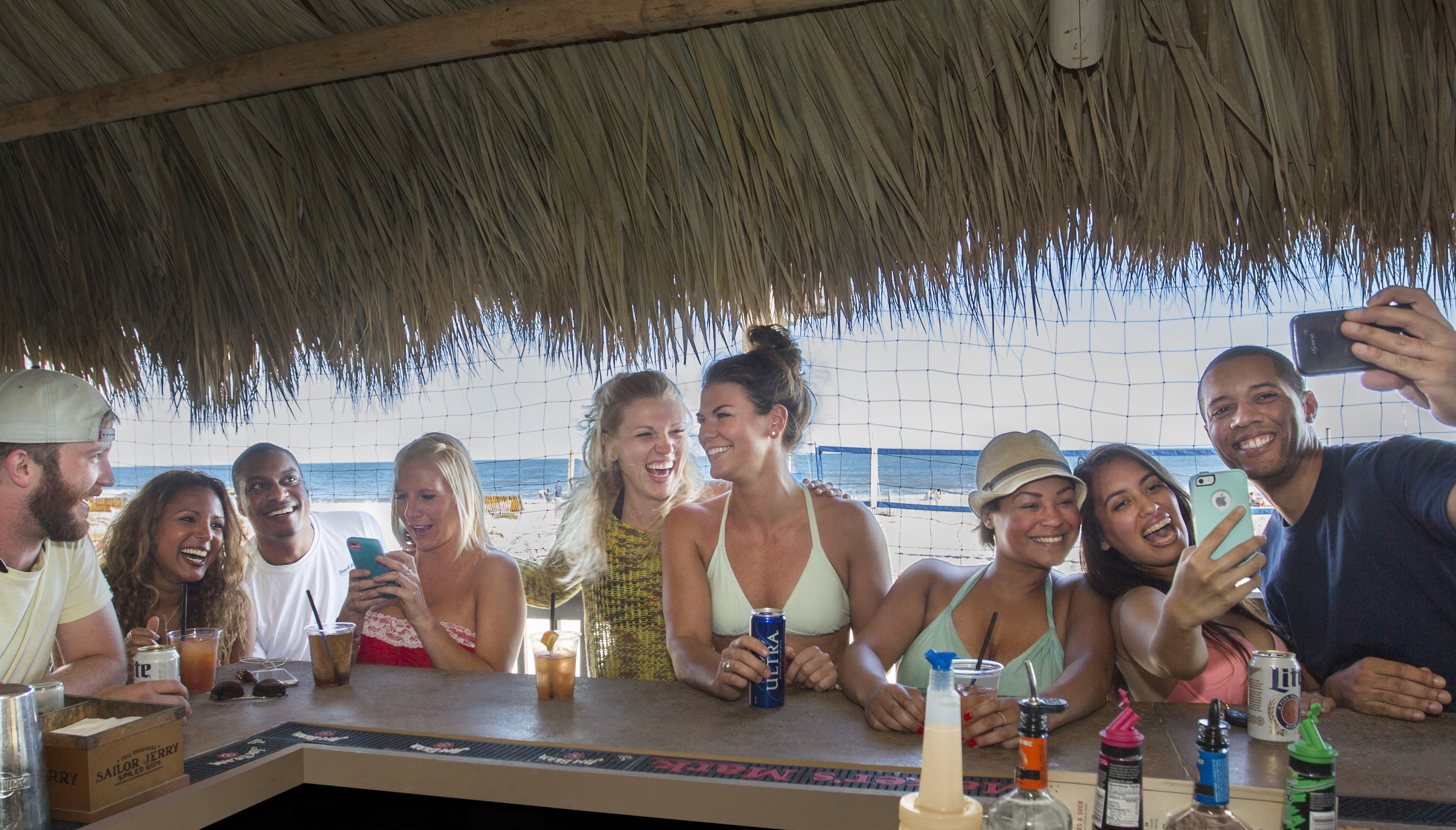 Un grupo de personas en un bar a la orilla de la playa con techo de paja, con vista al océano.