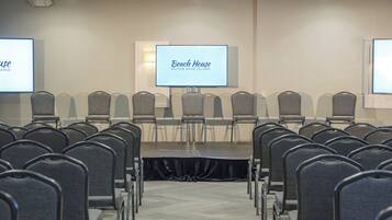 Uma sala de conferências com fileiras de cadeiras cinzas, um palco e uma tela exibindo "Beach House Hilton Head Island".