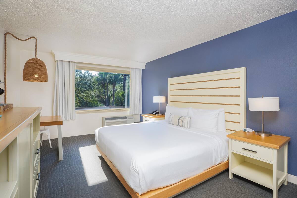 A hotel room with a large bed, bedside tables, a desk, and a window with a view of trees.