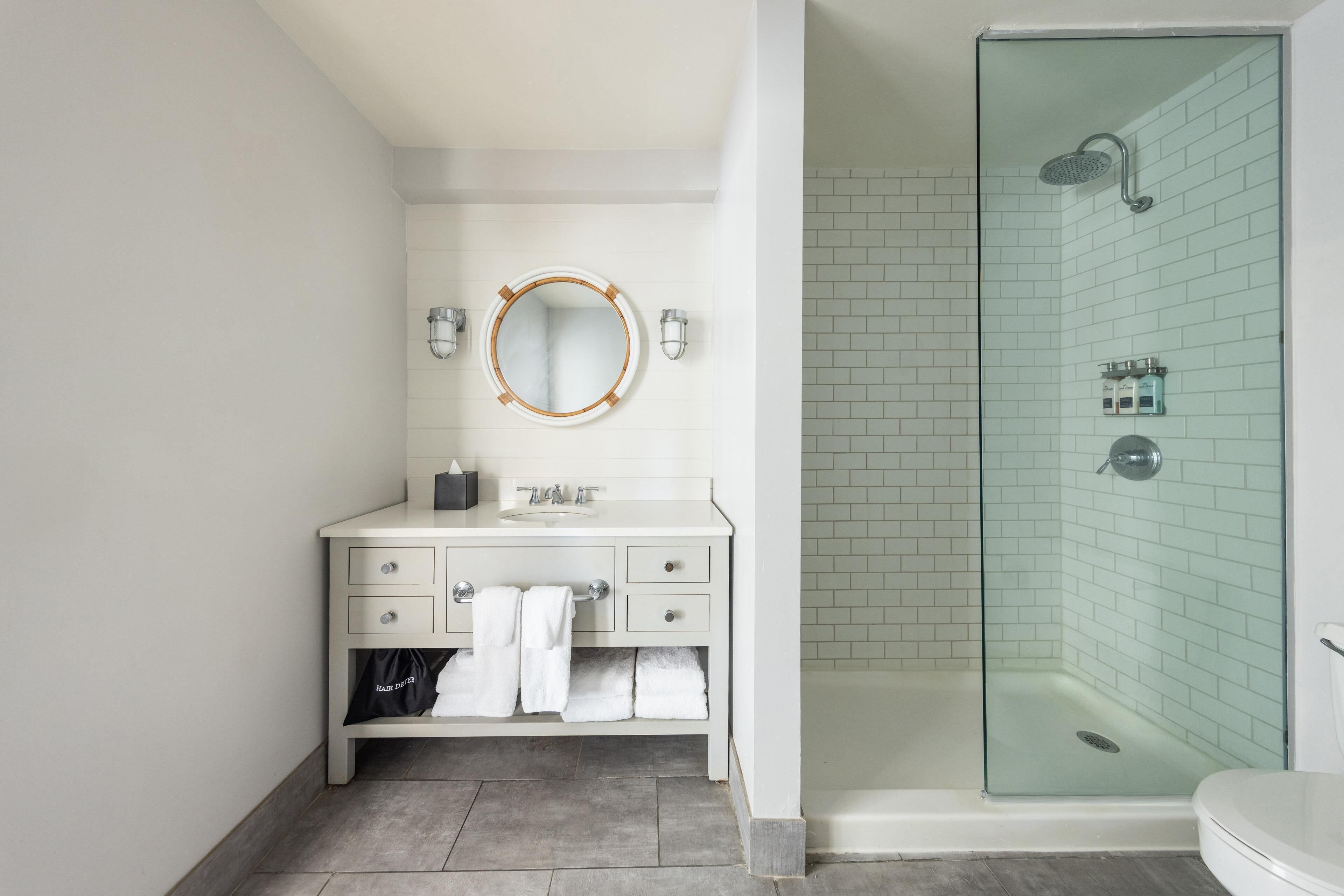 Une salle de bain avec un meuble-lavabo blanc, un miroir rond, une douche à parois de verre et des toilettes.