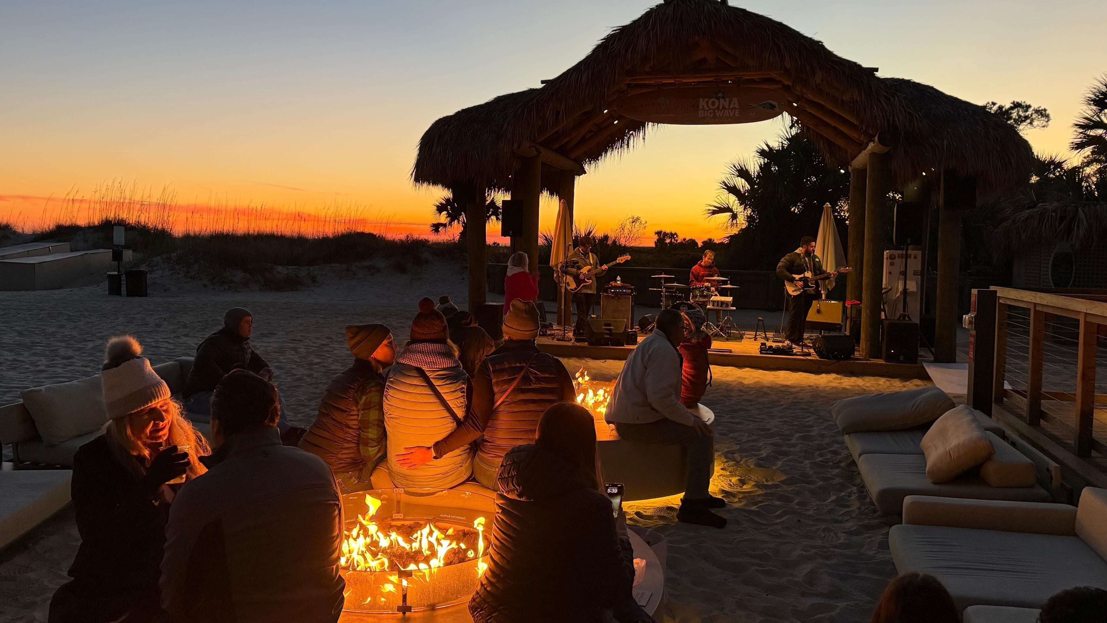 A beachside event with a stage, seated audience, and a fire pit.