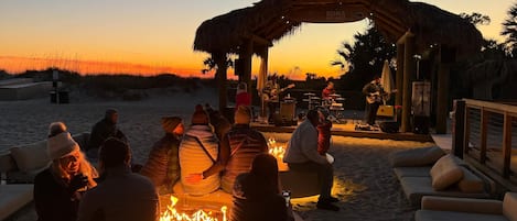 A beachside event with a stage, seated audience, and a fire pit.