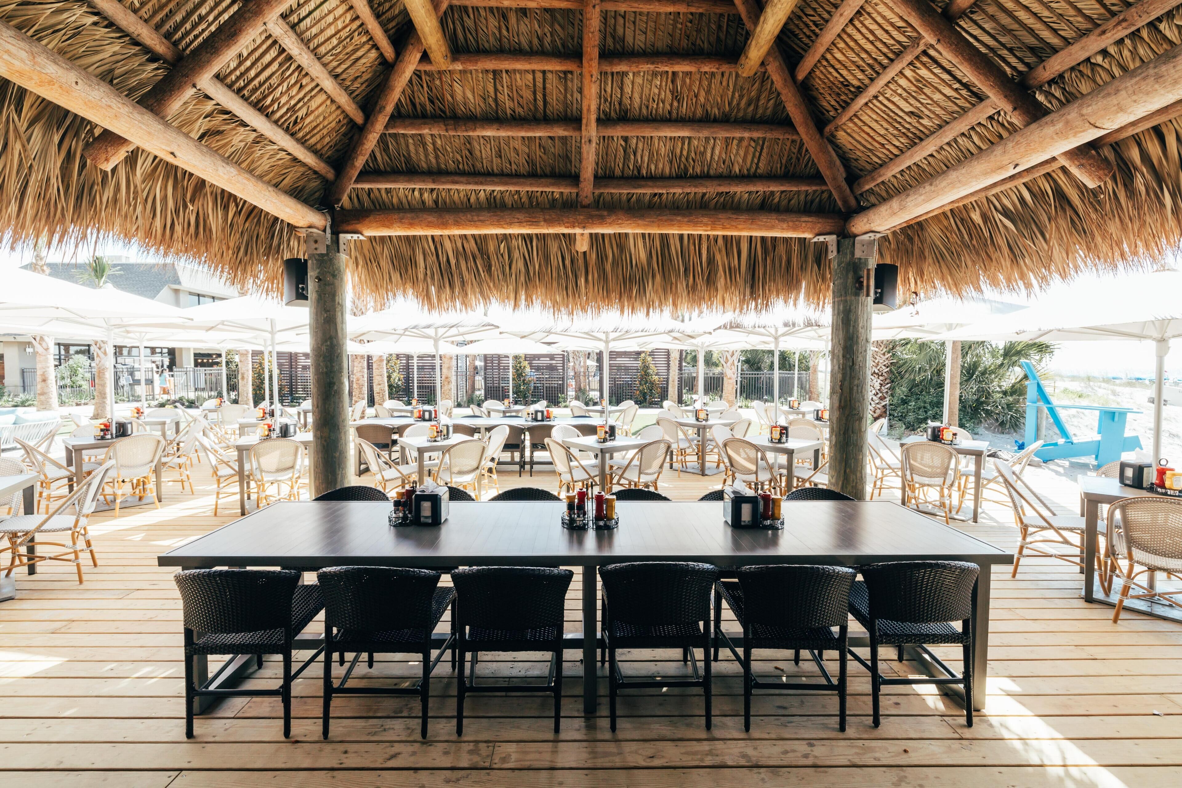 An outdoor dining area with a thatched roof, wooden tables, and wicker chairs.
