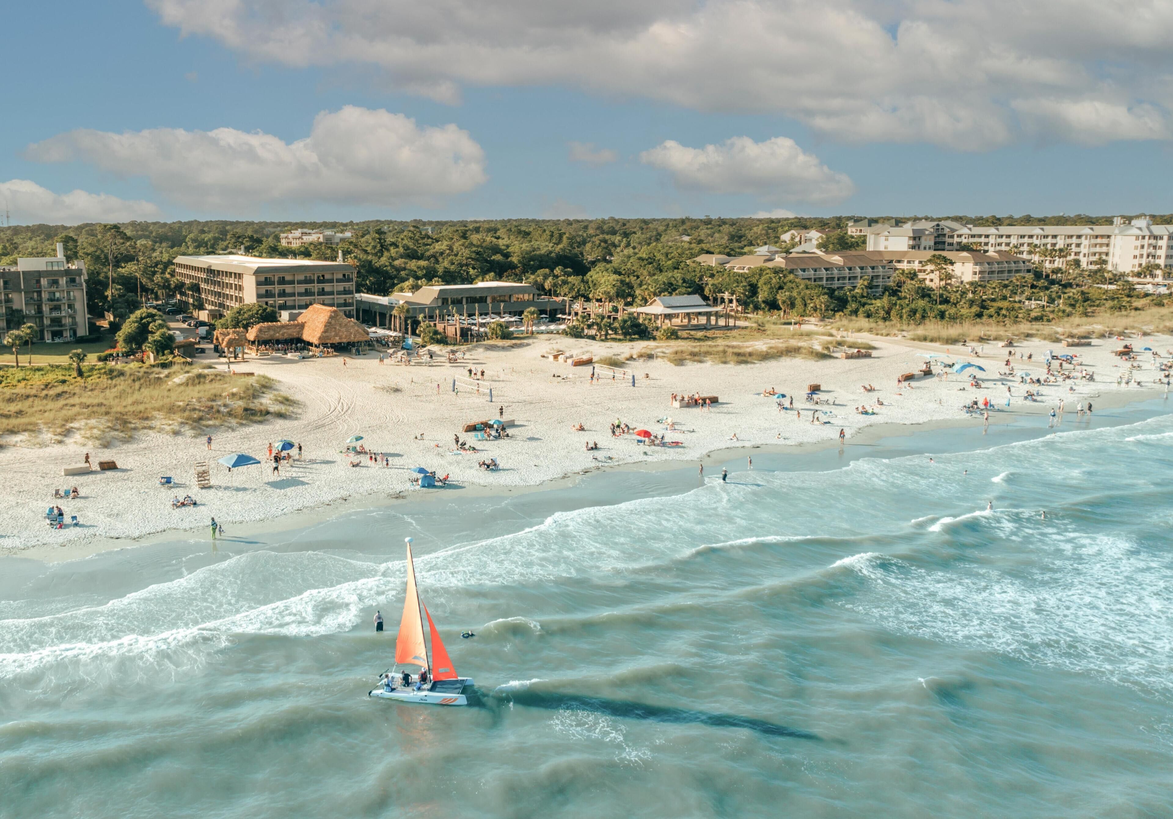 A beach with a sailboat, sandy shore, and buildings in the background.