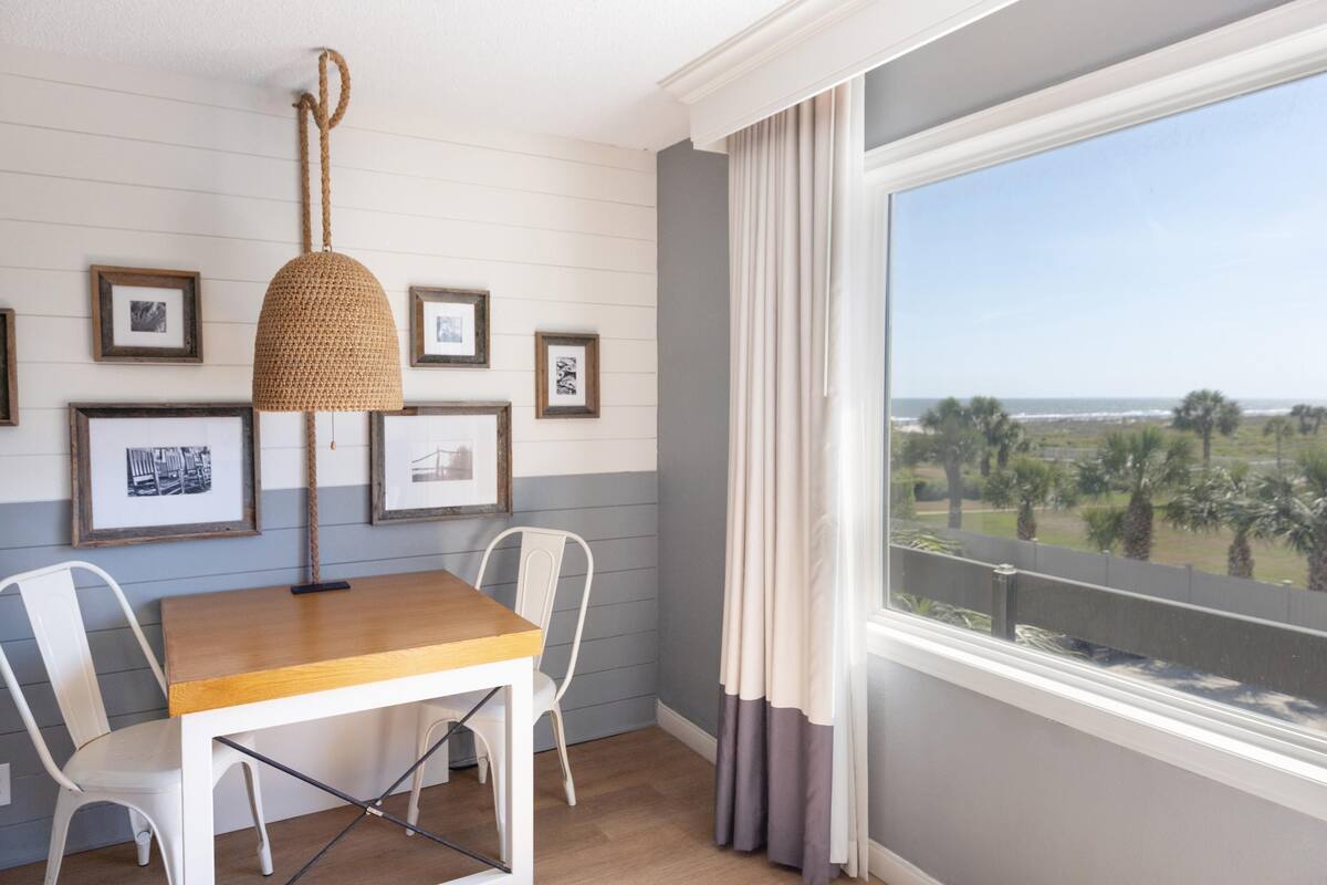 A room with a wooden table, white chairs, a woven pendant light, framed pictures on the wall, and a view of the ocean through a window with curtains.