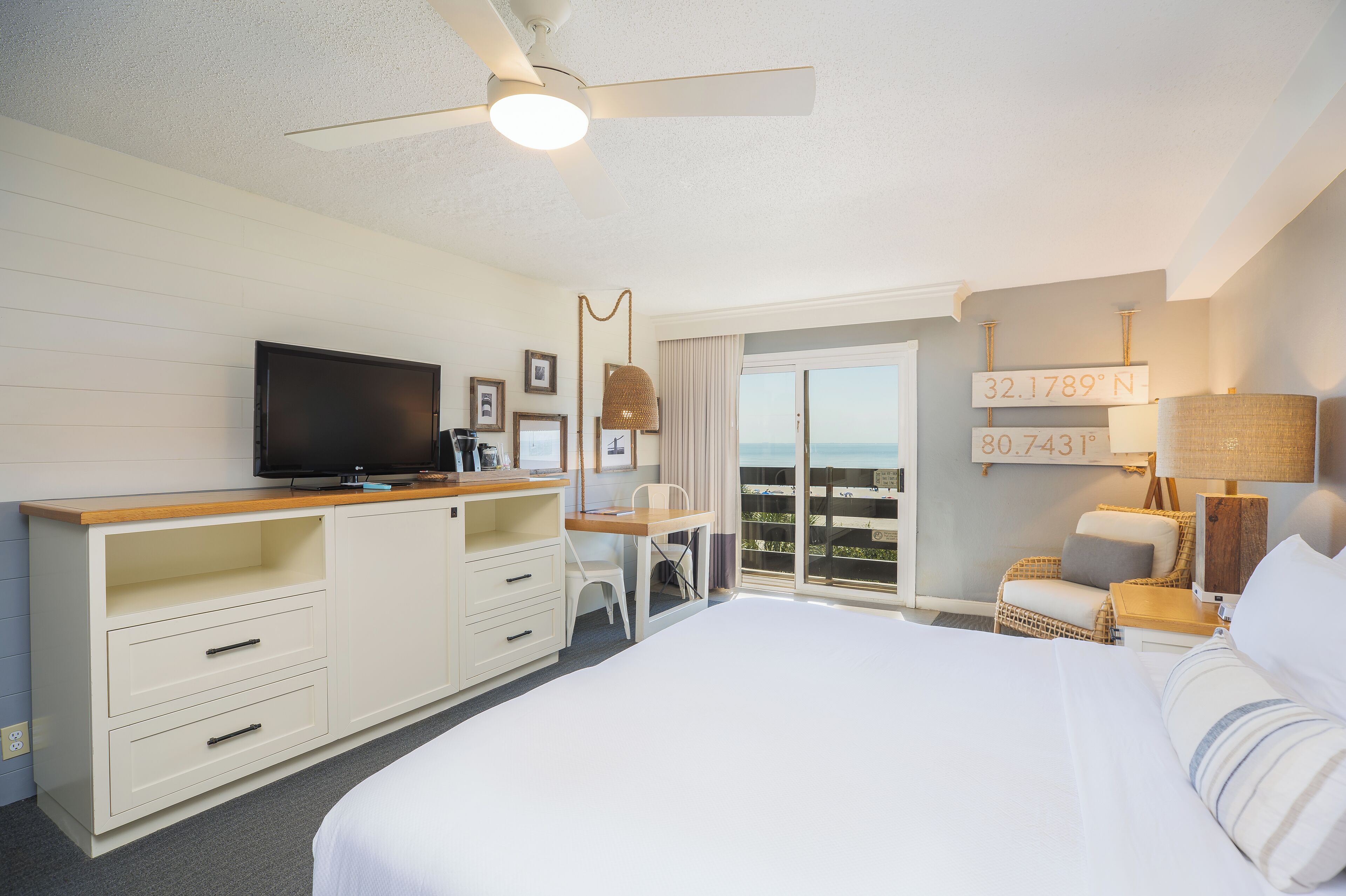 A hotel room with a bed, a desk, a television, a ceiling fan, and a view of the ocean.