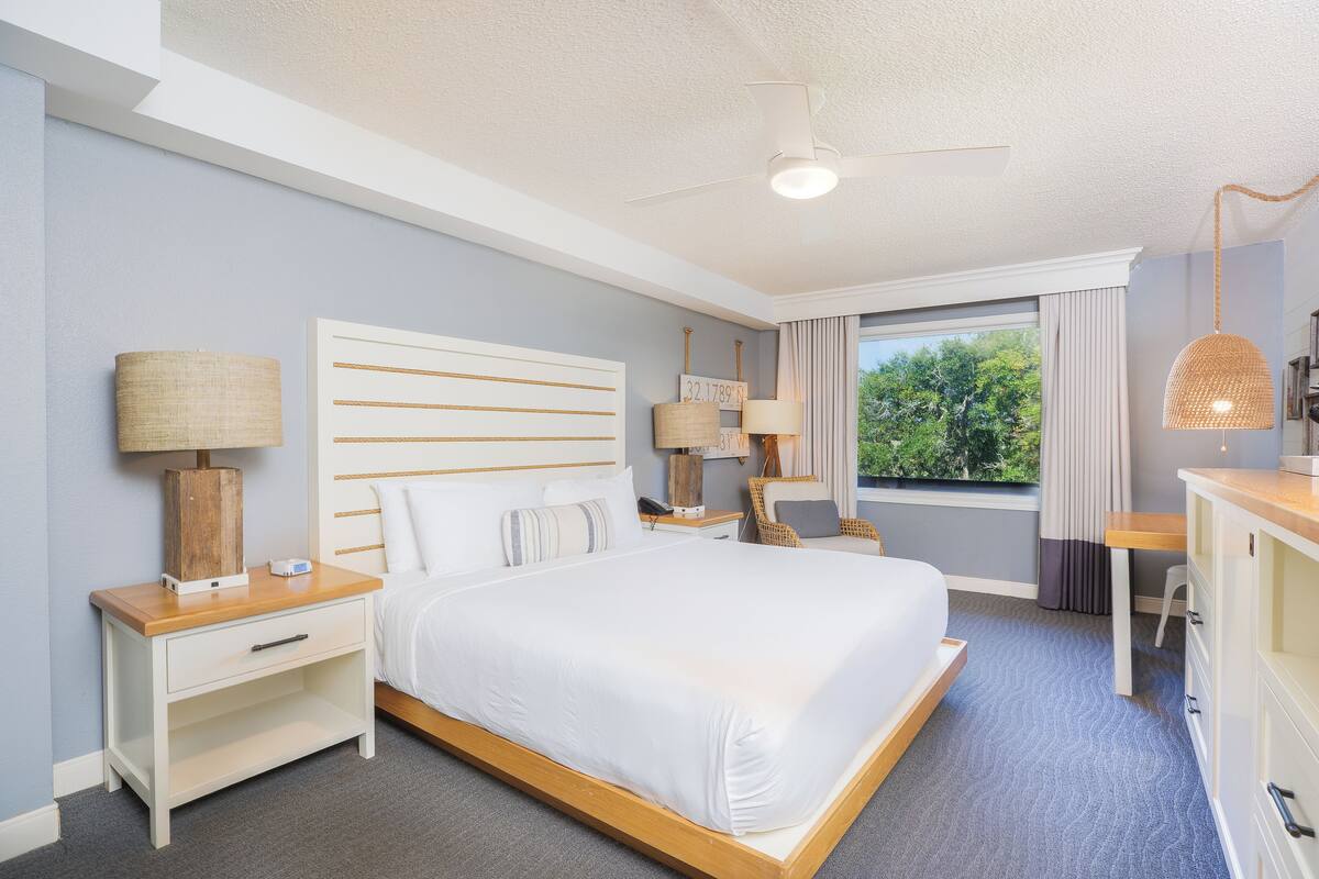 A modern hotel room with a large bed, bedside tables, a desk, and a view of the outdoors.