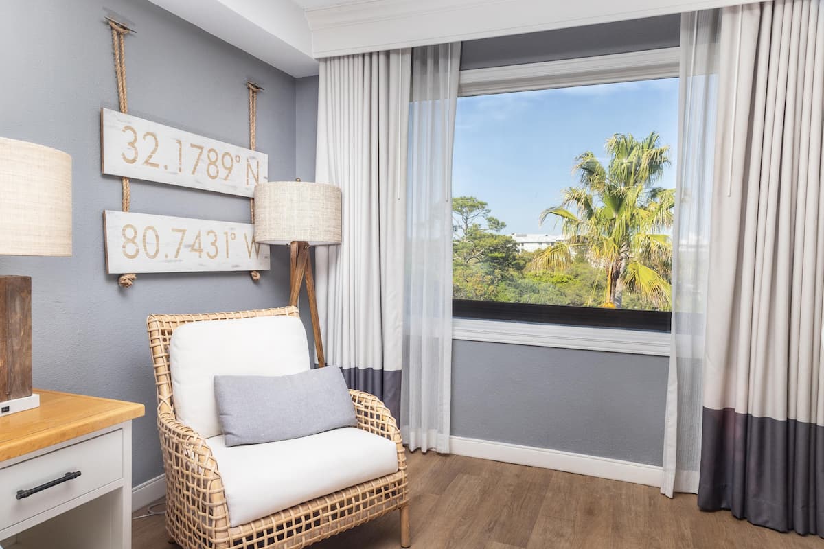 A room with a wicker chair, a nightstand, a lamp, and a window with curtains.