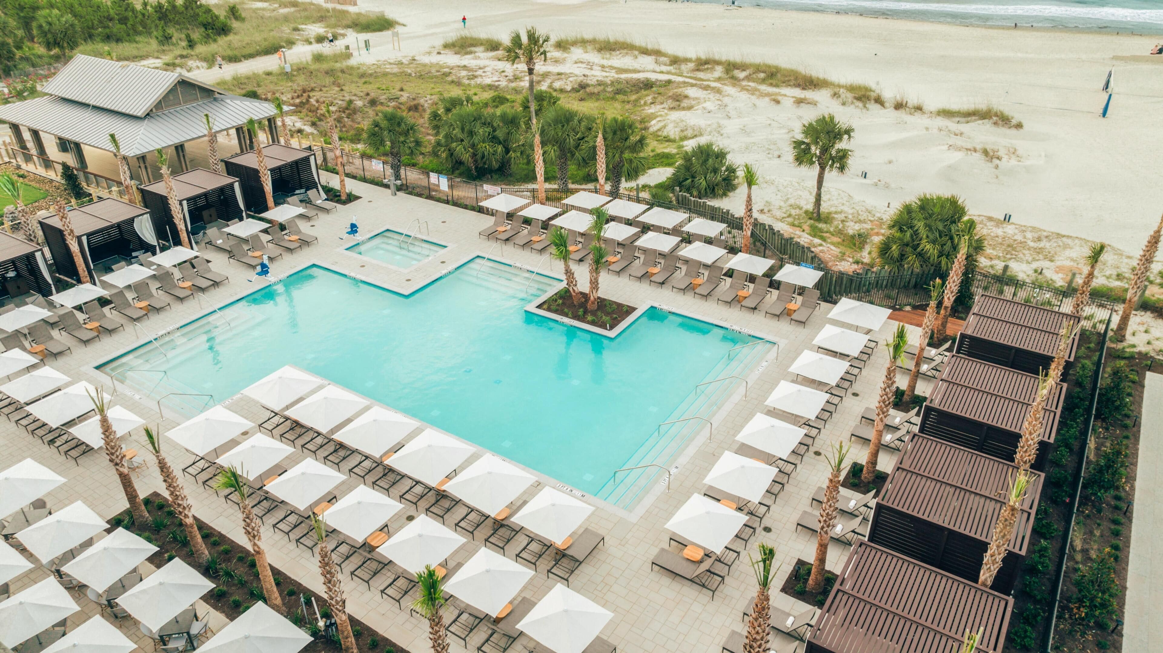 Un espace piscine doté de chaises longues et de parasols, entouré de palmiers et avec une plage en arrière-plan.