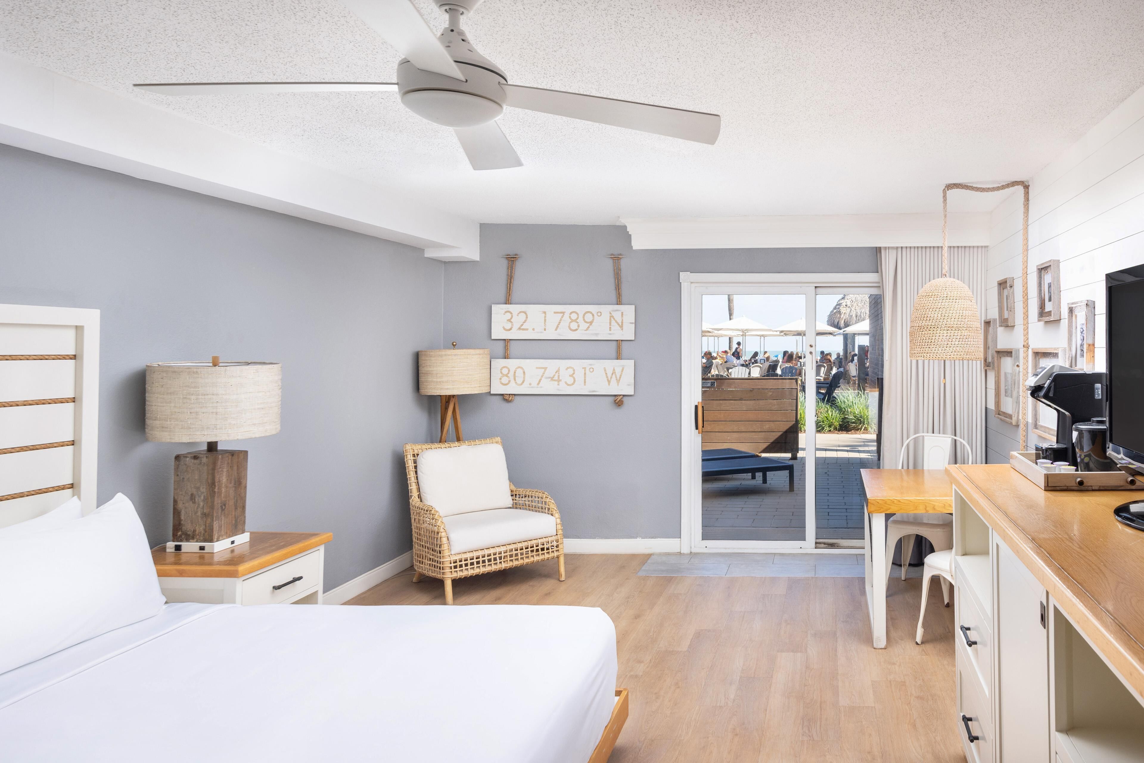A hotel room with a bed, a chair, a desk, a ceiling fan, and a view through a glass door.