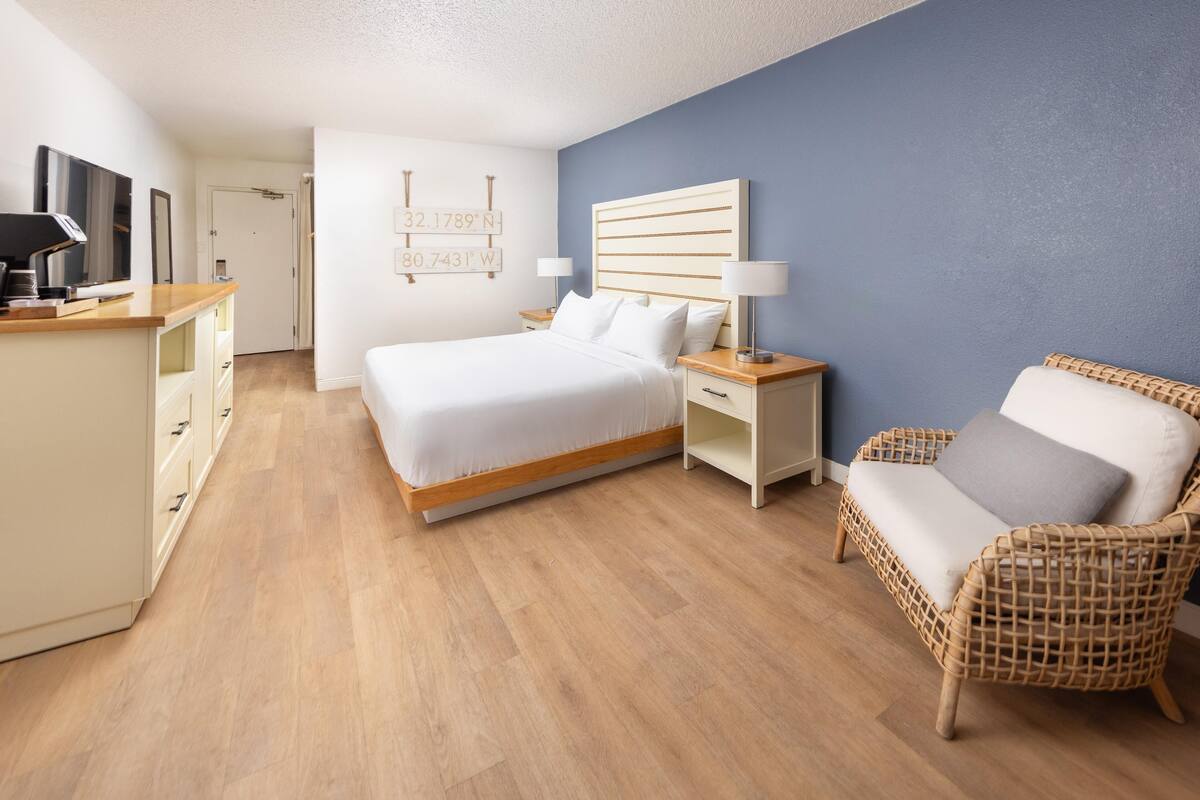 A hotel room with a bed, bedside tables, a TV, a wicker chair, and a wall with coordinates.