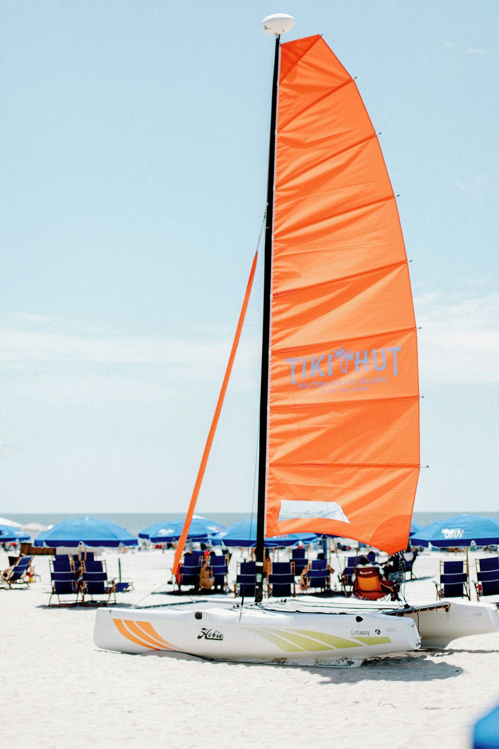 Ein Segelboot mit orangefarbenem Segel an einem sandigen Strand, im Hintergrund sind blaue Sonnenschirme zu sehen.