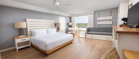 A modern hotel room with a bed, bedside tables, a desk, a chair, a TV, and a view of the outdoors.