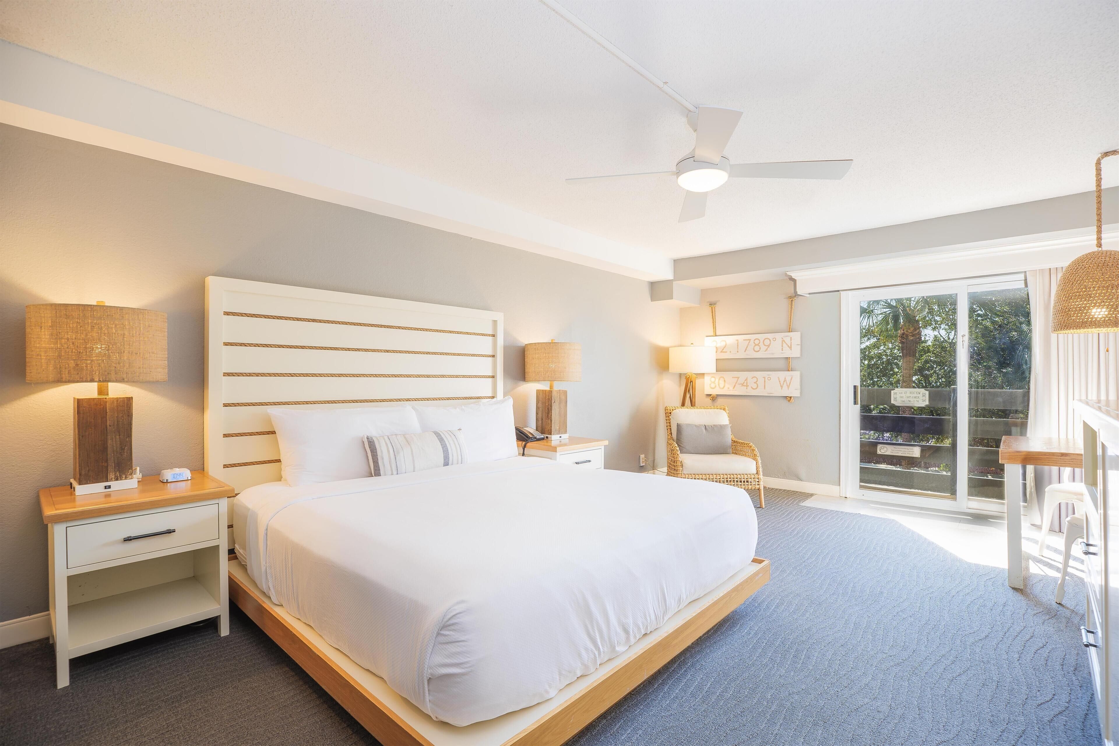 A modern bedroom with a large bed, two bedside tables, a ceiling fan, and a view of the outdoors through a sliding glass door.