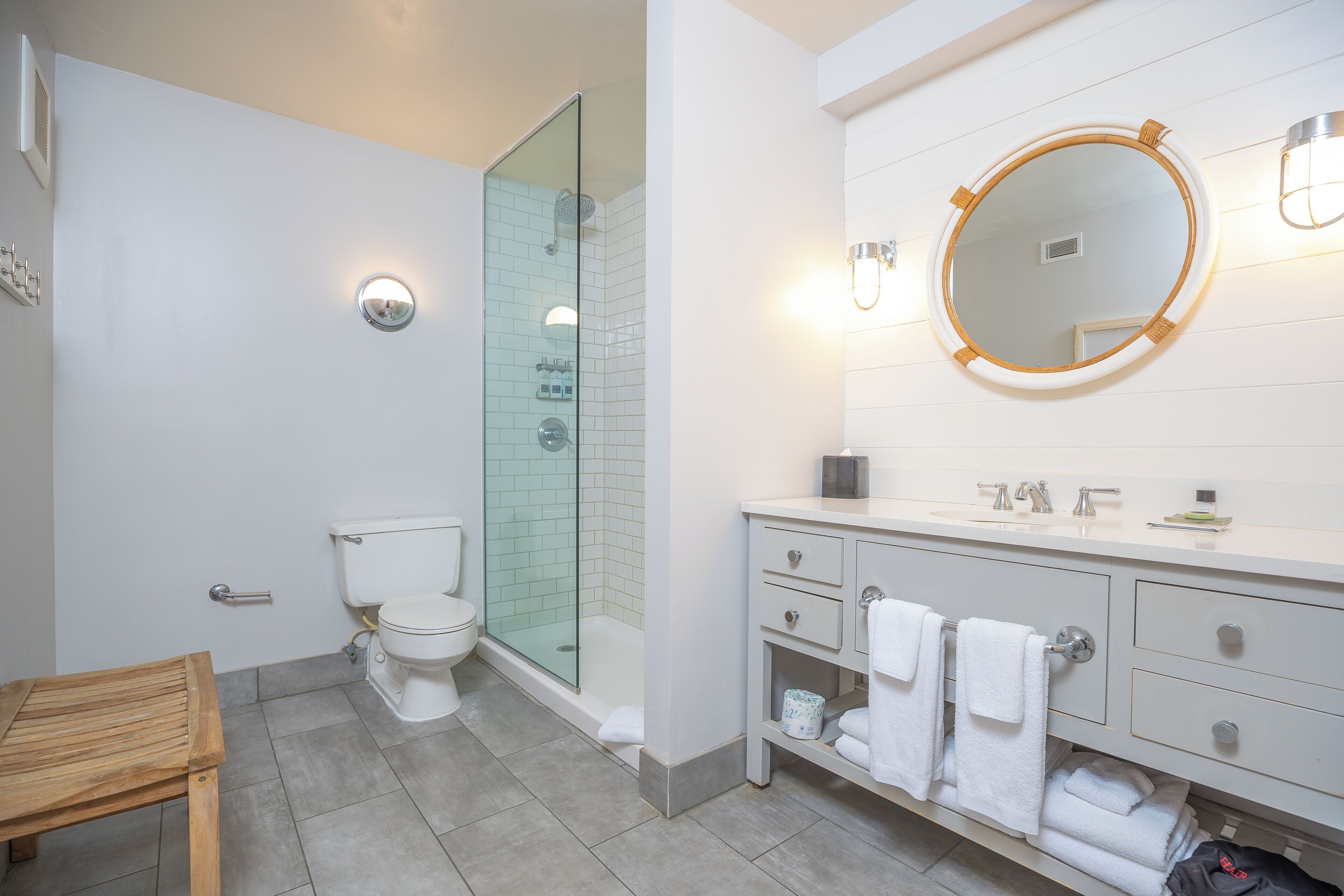 Une salle de bain moderne avec une douche à parois de verre, un lavabo blanc avec un miroir rond et un banc en bois.