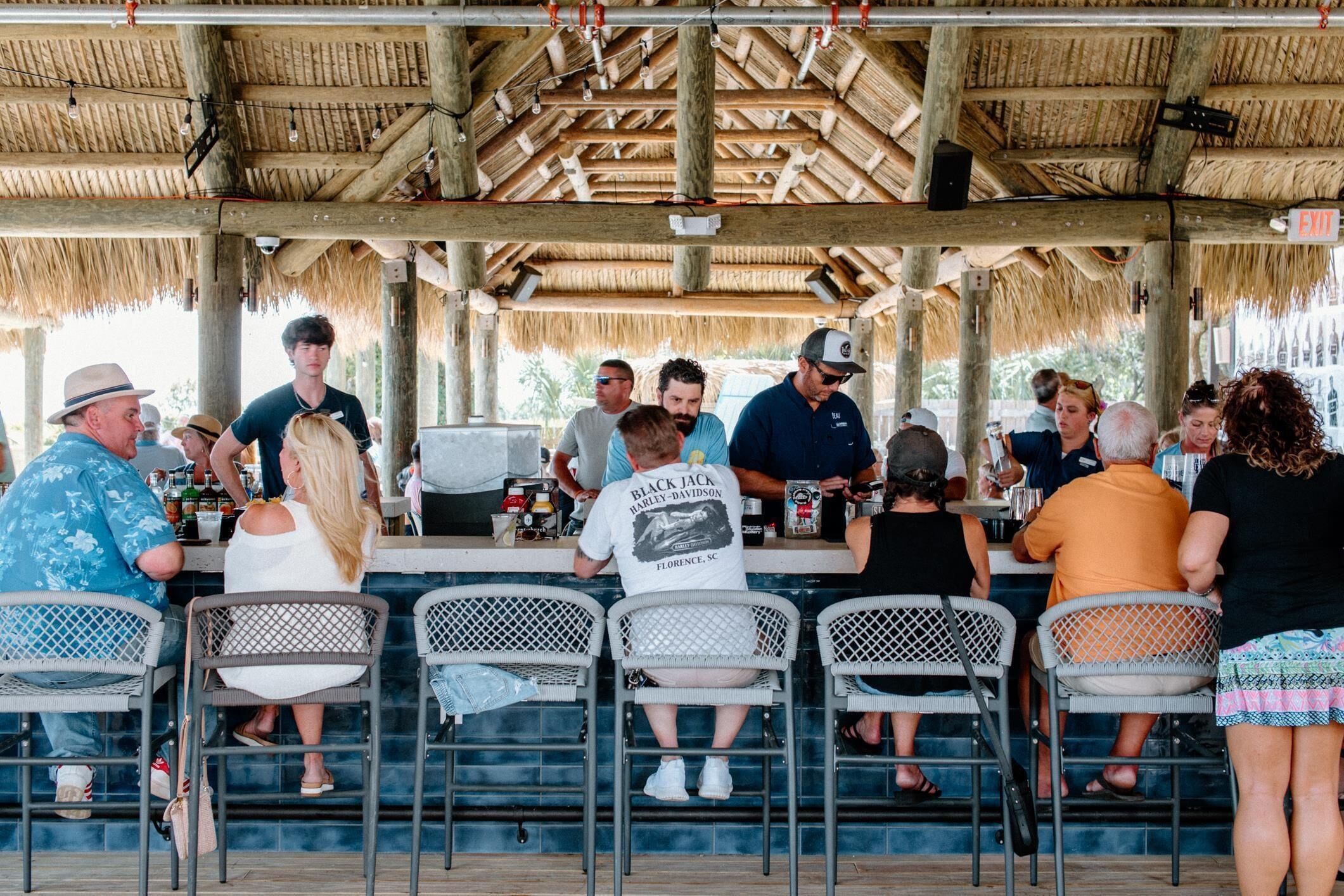 A covered outdoor bar area with metal chairs and tables, a thatched roof, and a visible exit sign.