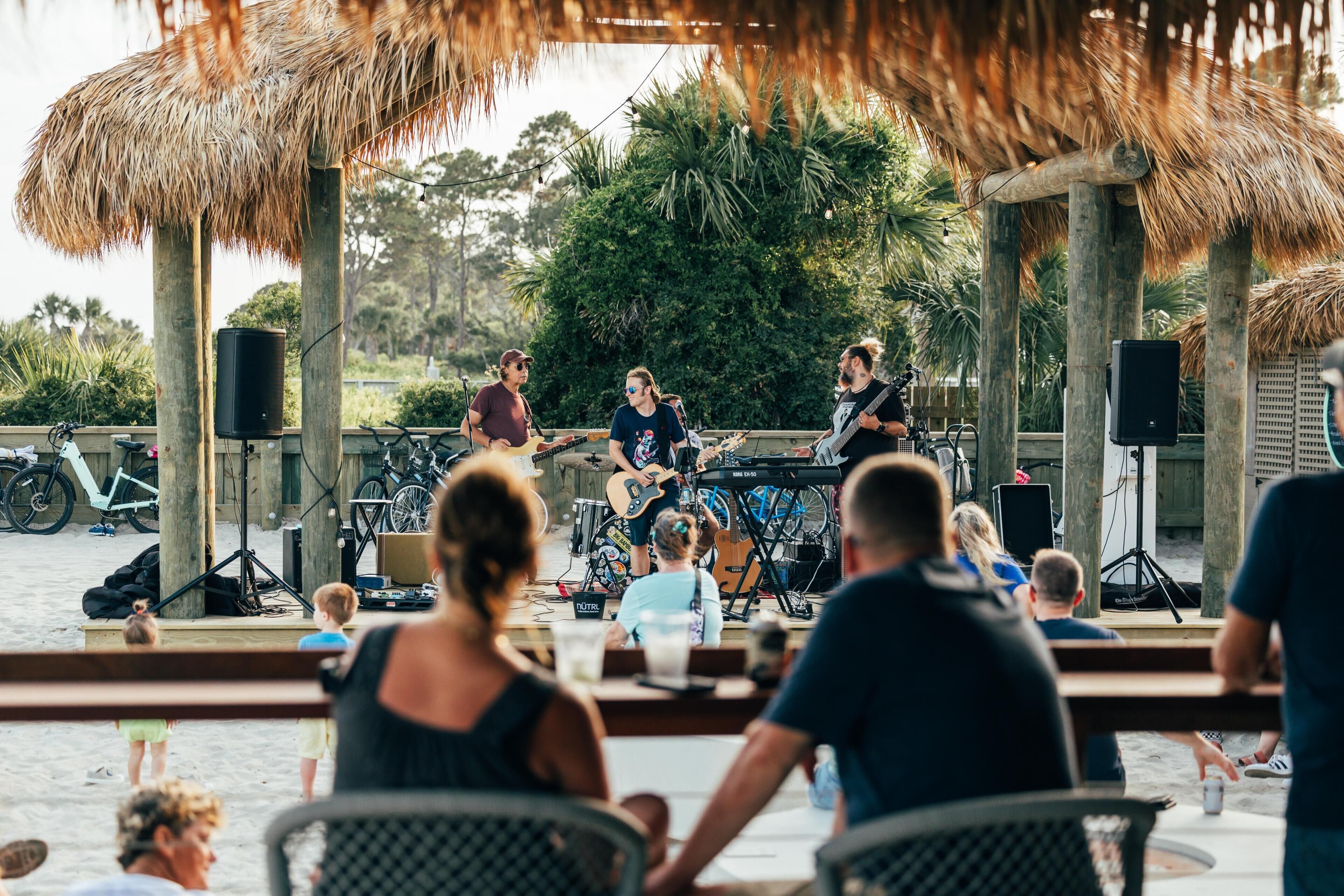 Une scène installée sur la plage, où des musiciens se produisent, entourée de personnes qui profitent de la plage.