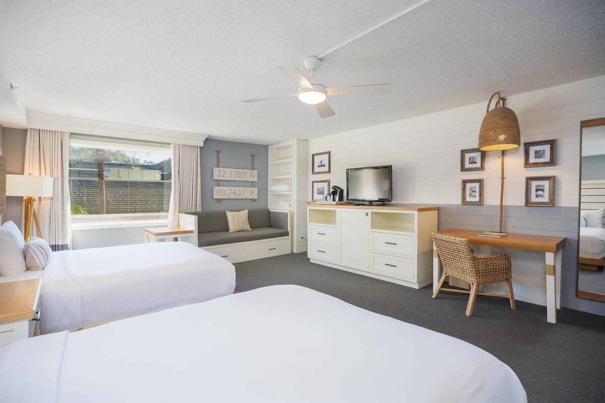 A hotel room with two beds, a desk, a television, and a ceiling fan.