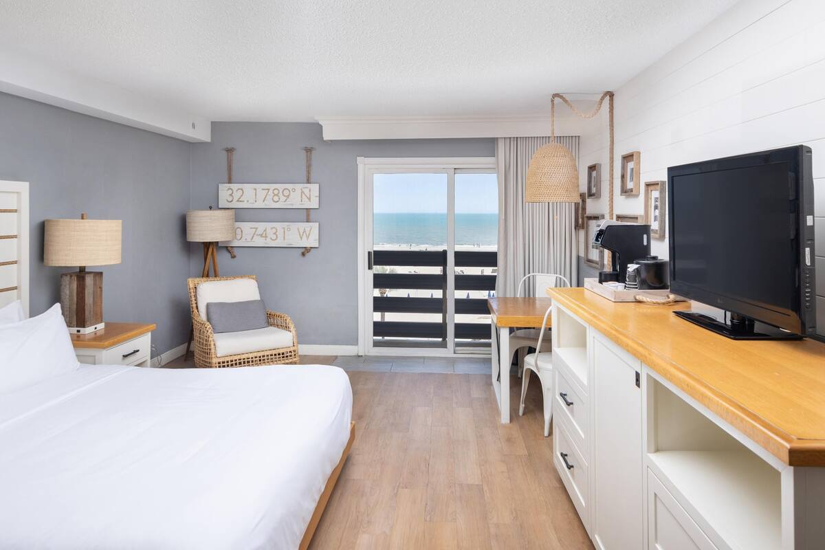 Hotelkamer met een bed, een stoel, een bureau met een televisie, een koffiezetapparaat en uitzicht op het strand.