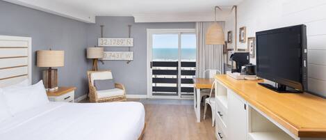 Une chambre d’hôtel avec un lit, une chaise, un bureau avec une télévision, une machine à café et une vue sur la plage.