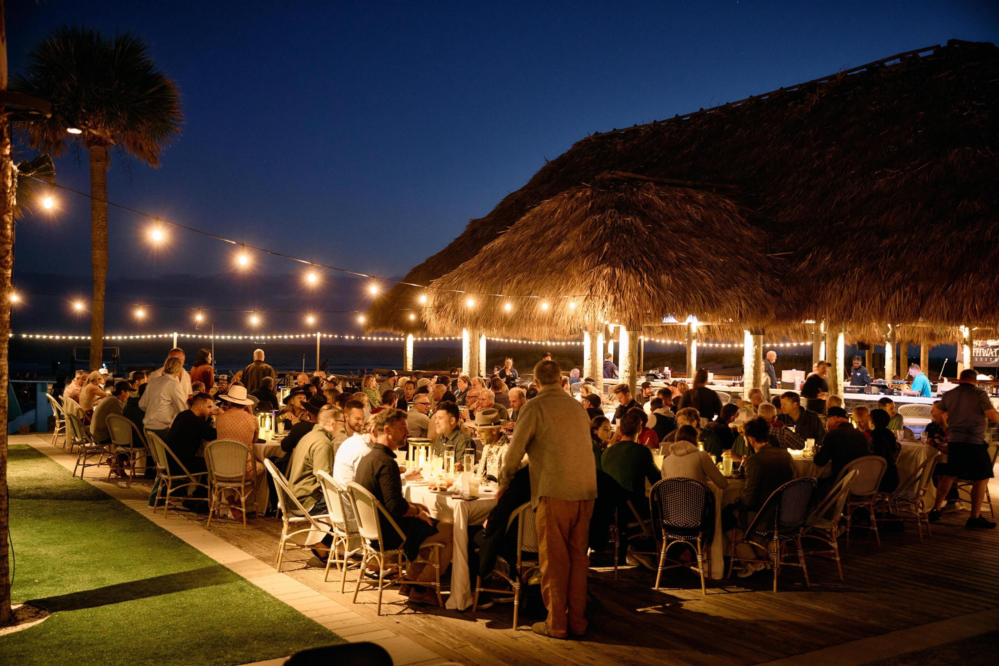 An outdoor dining area with tables and chairs, a thatched-roof structure, and string lights.
