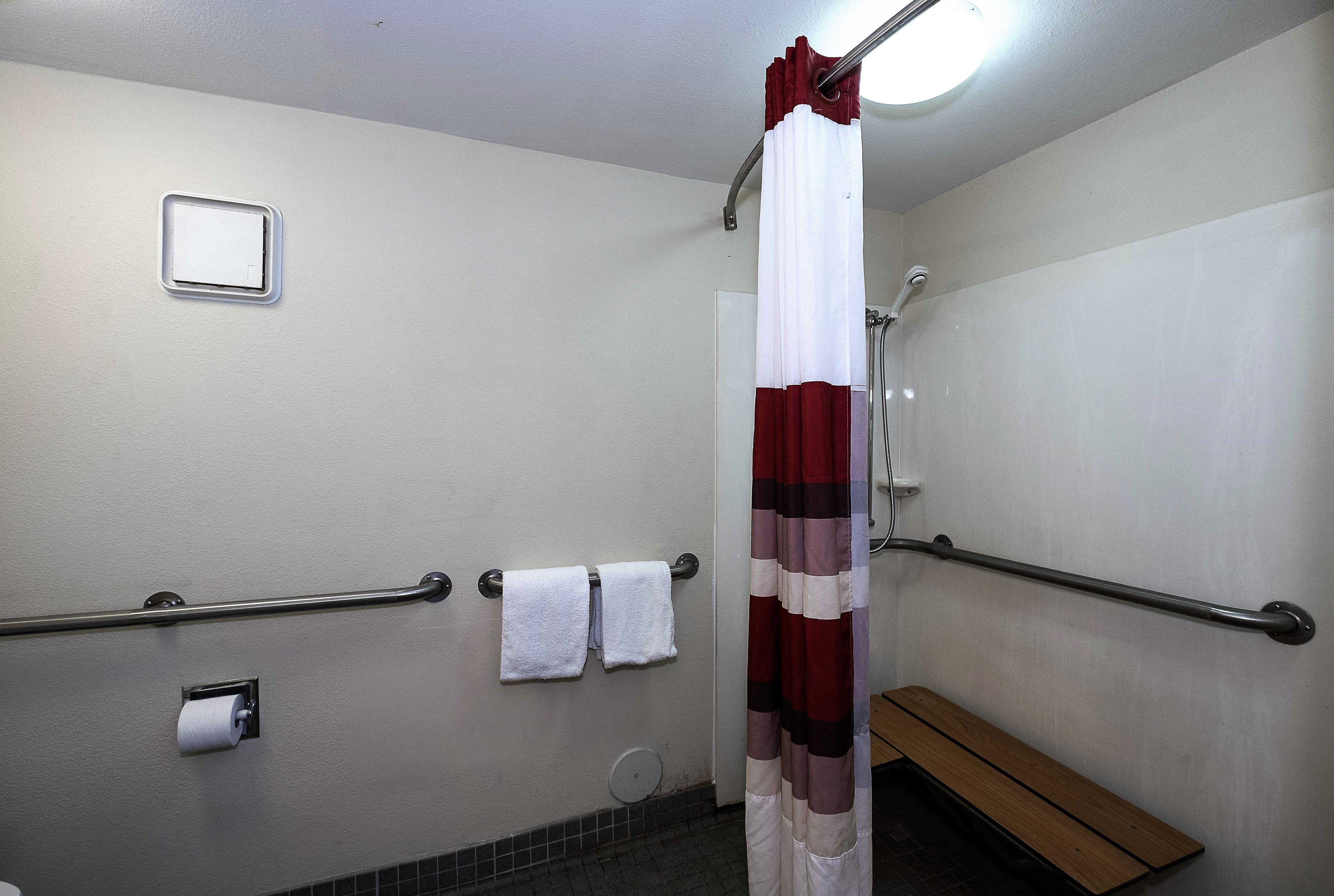 Un baño con cortina de ducha, barras de apoyo, un banco y un dispensador de papel higiénico.