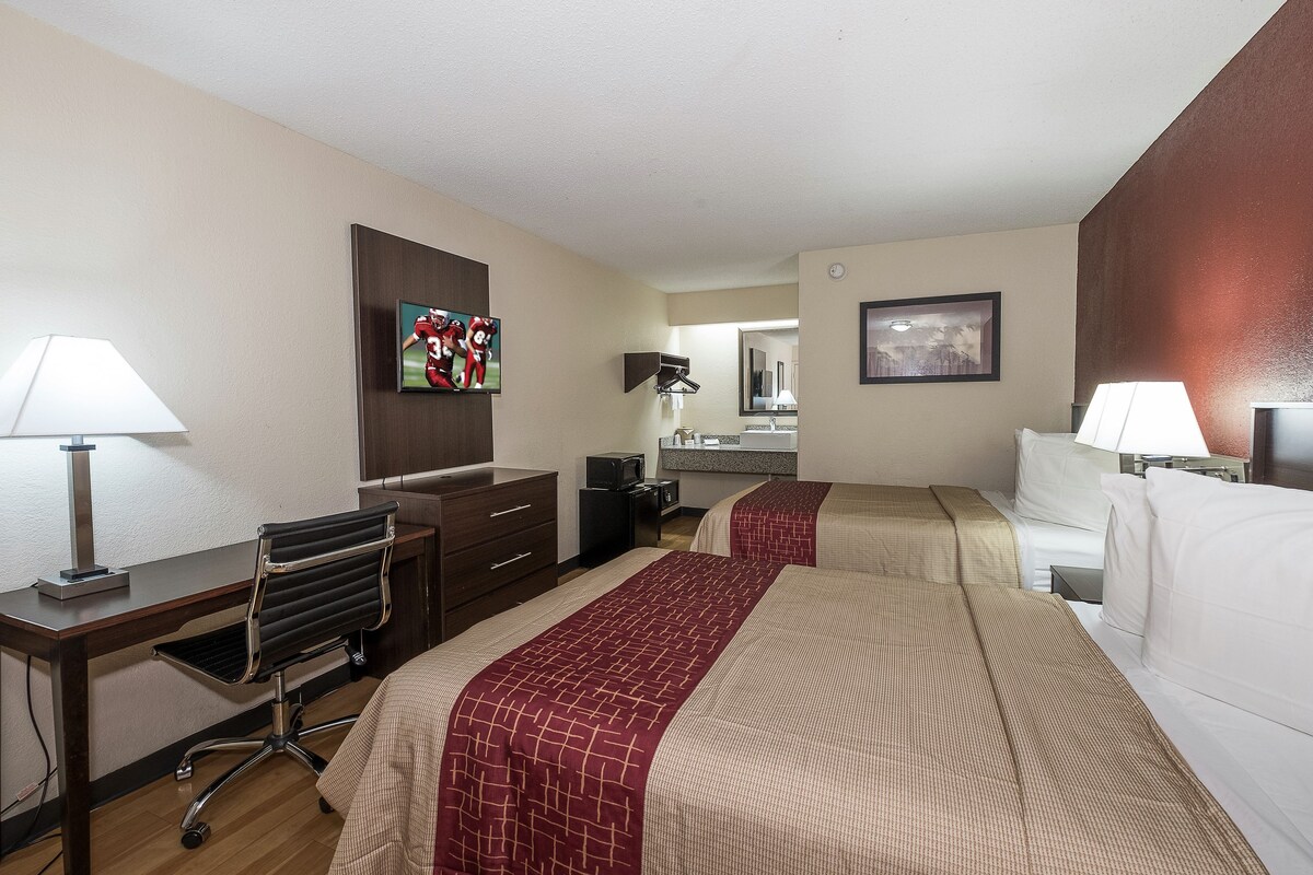A hotel room with two beds, a desk, a chair, a television, a mirror, and a bathroom.