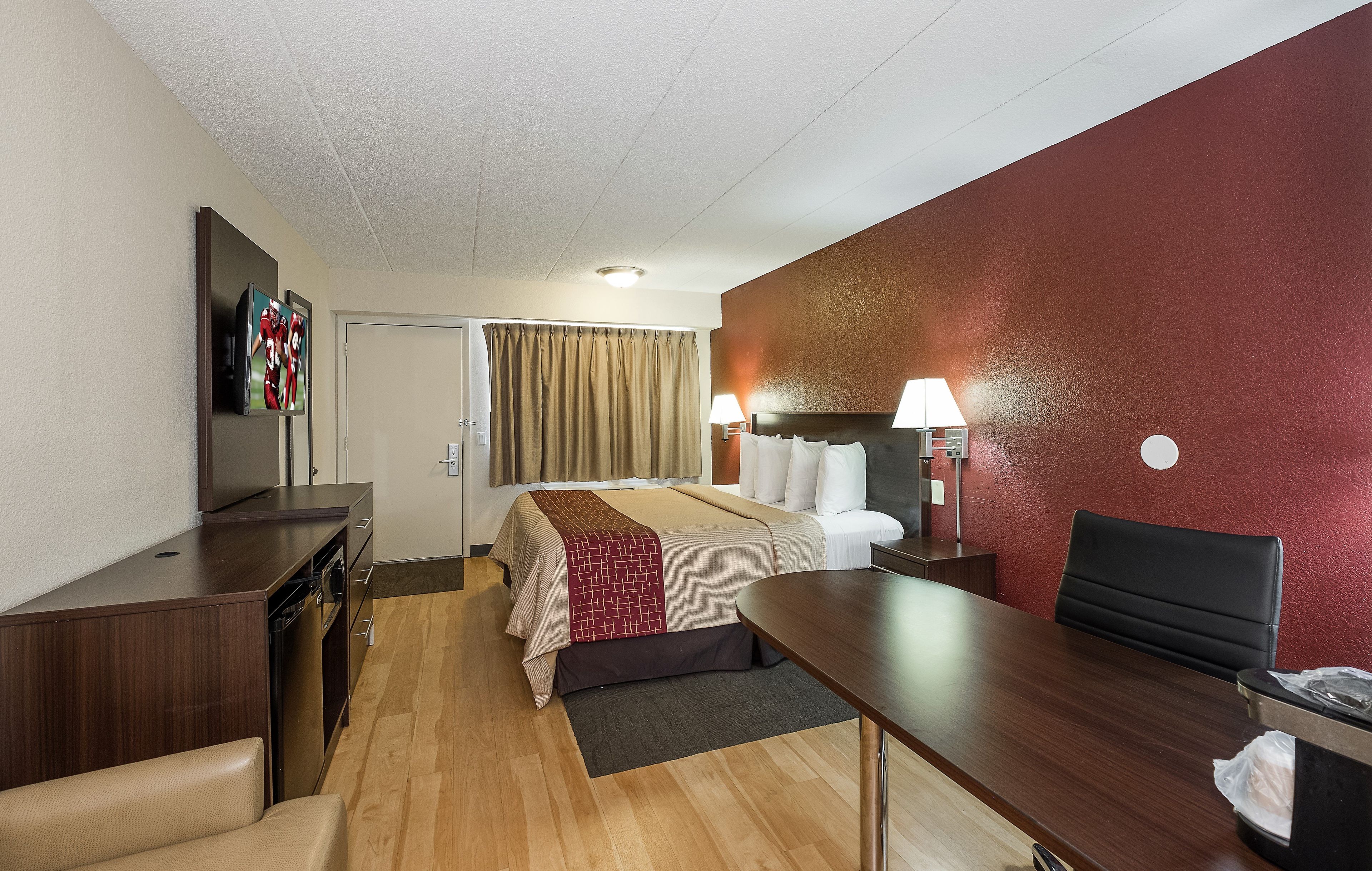 A hotel room with a bed, desk, chair, TV, and a red accent wall.