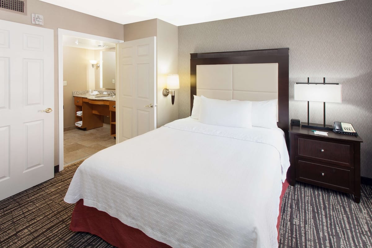 A hotel room with a large bed, a bedside table with a lamp, a desk with a telephone, and a bathroom visible through an open door.