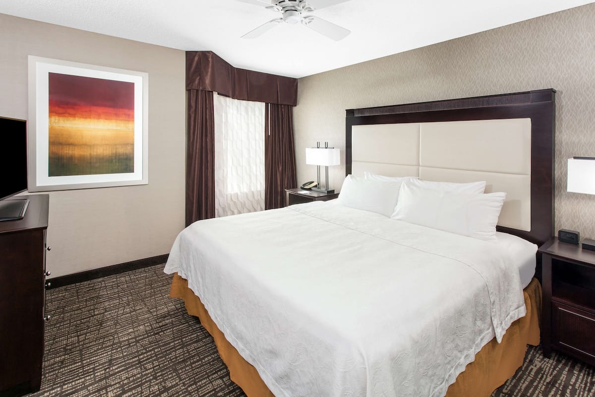 A hotel room with a large bed, a ceiling fan, a nightstand with a lamp, a desk with a telephone, and a framed picture on the wall.