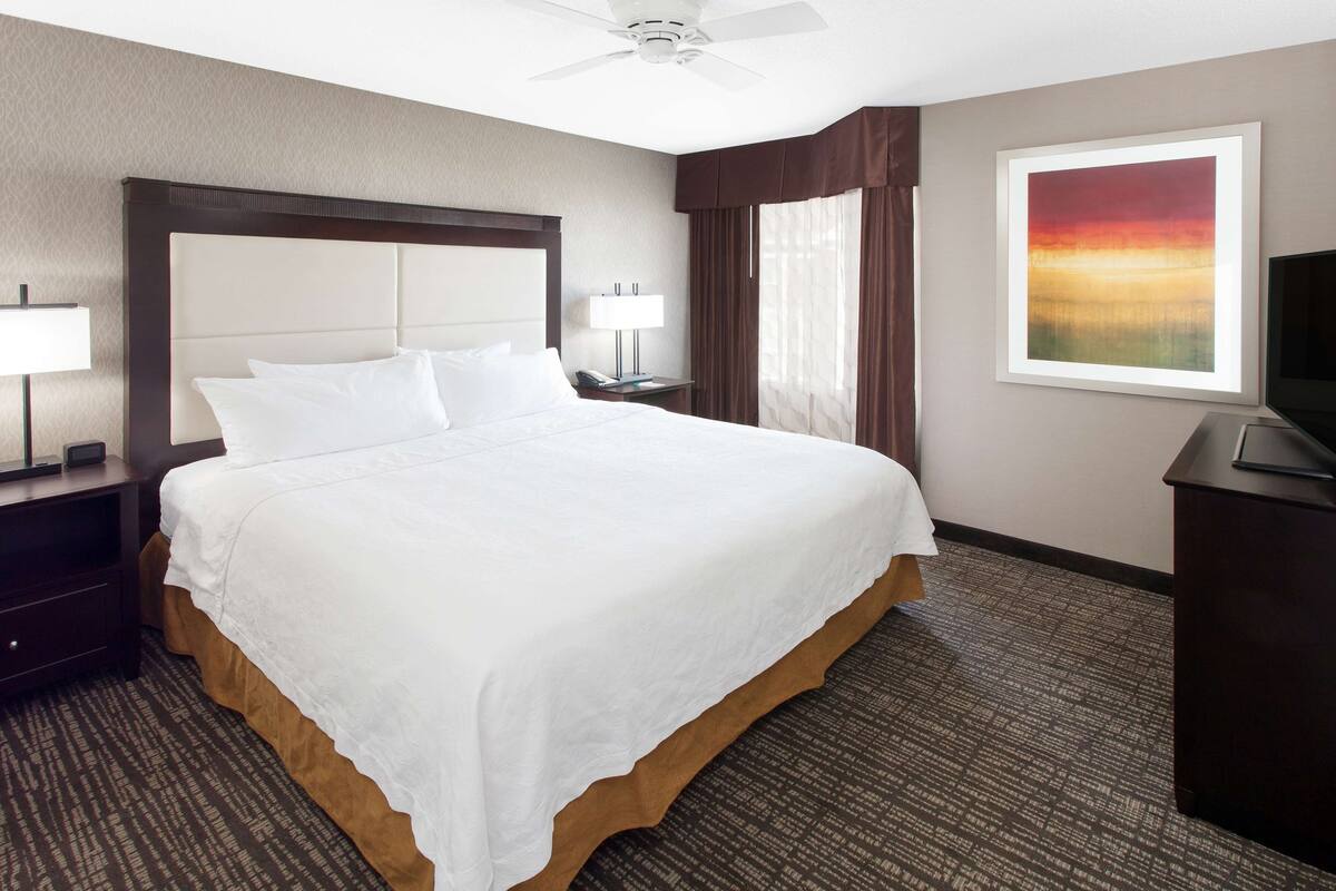 A hotel room with a large bed, a ceiling fan, a desk with a lamp, a TV, and a painting on the wall.