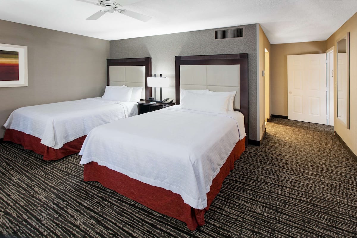A hotel room with two beds, a ceiling fan, a nightstand with a lamp, and a door.