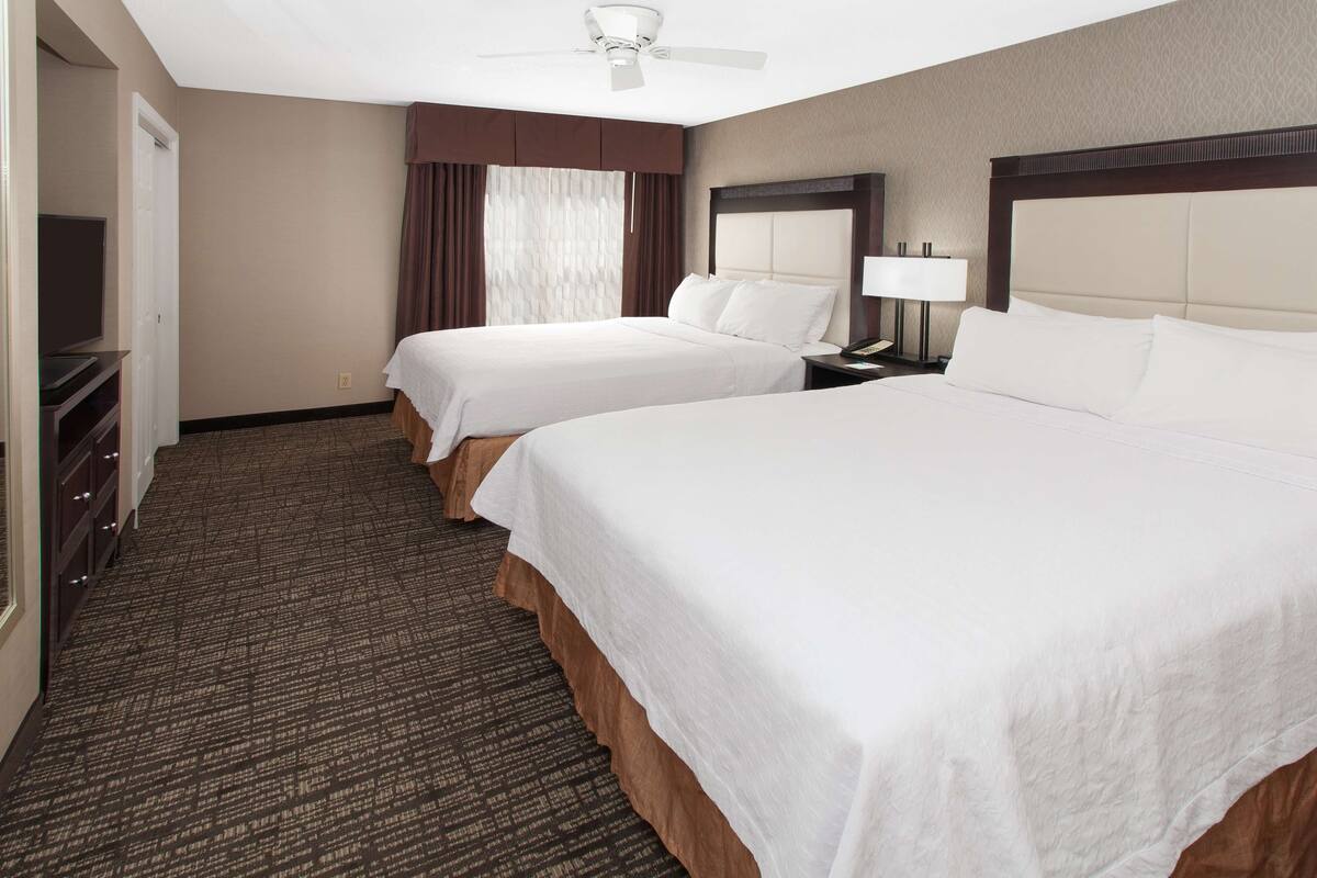 A hotel room with two beds, a ceiling fan, a TV, and a nightstand with a lamp.