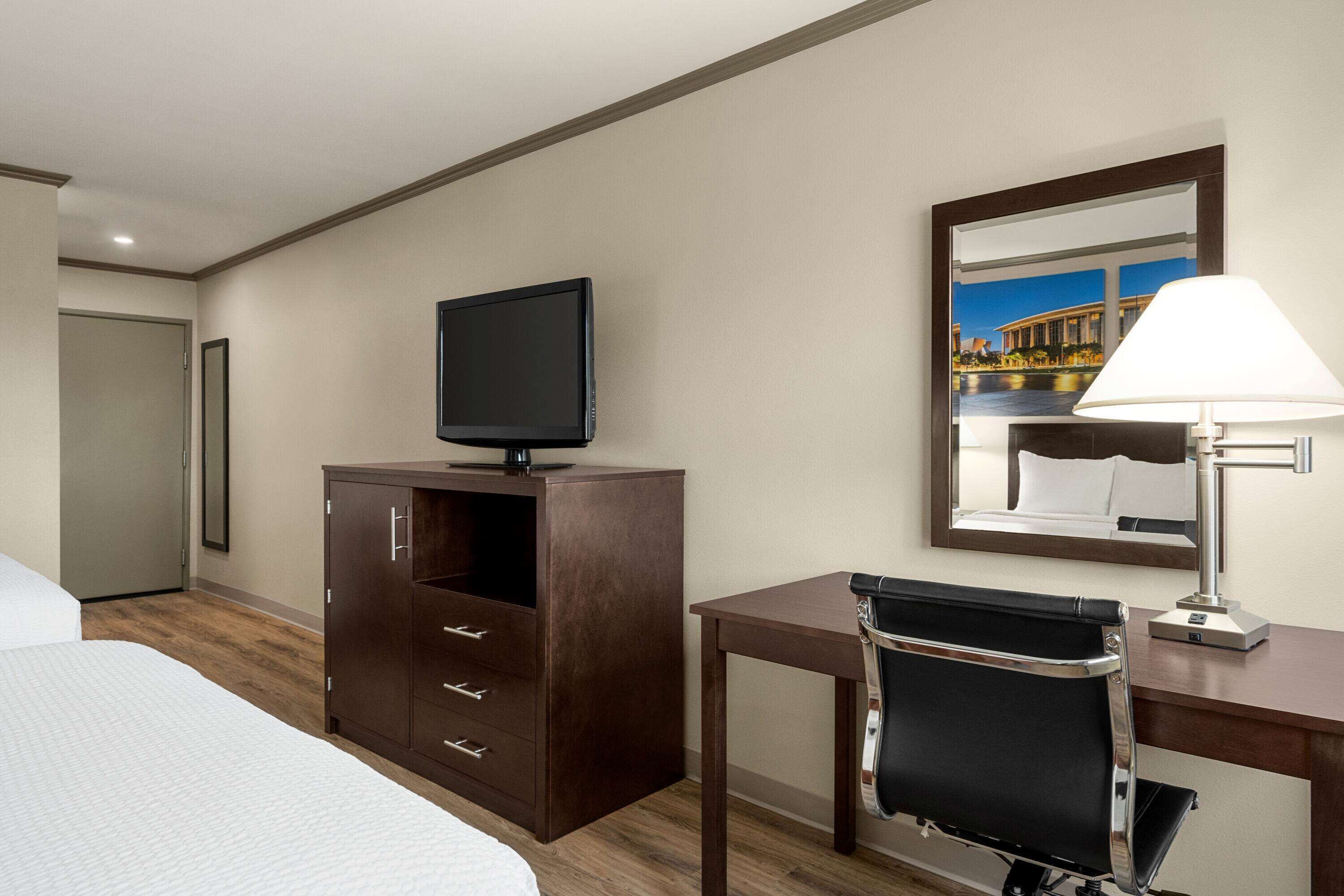 Habitación de hotel con cama, escritorio con silla, televisor en un mueble y espejo que refleja un paisaje urbano.